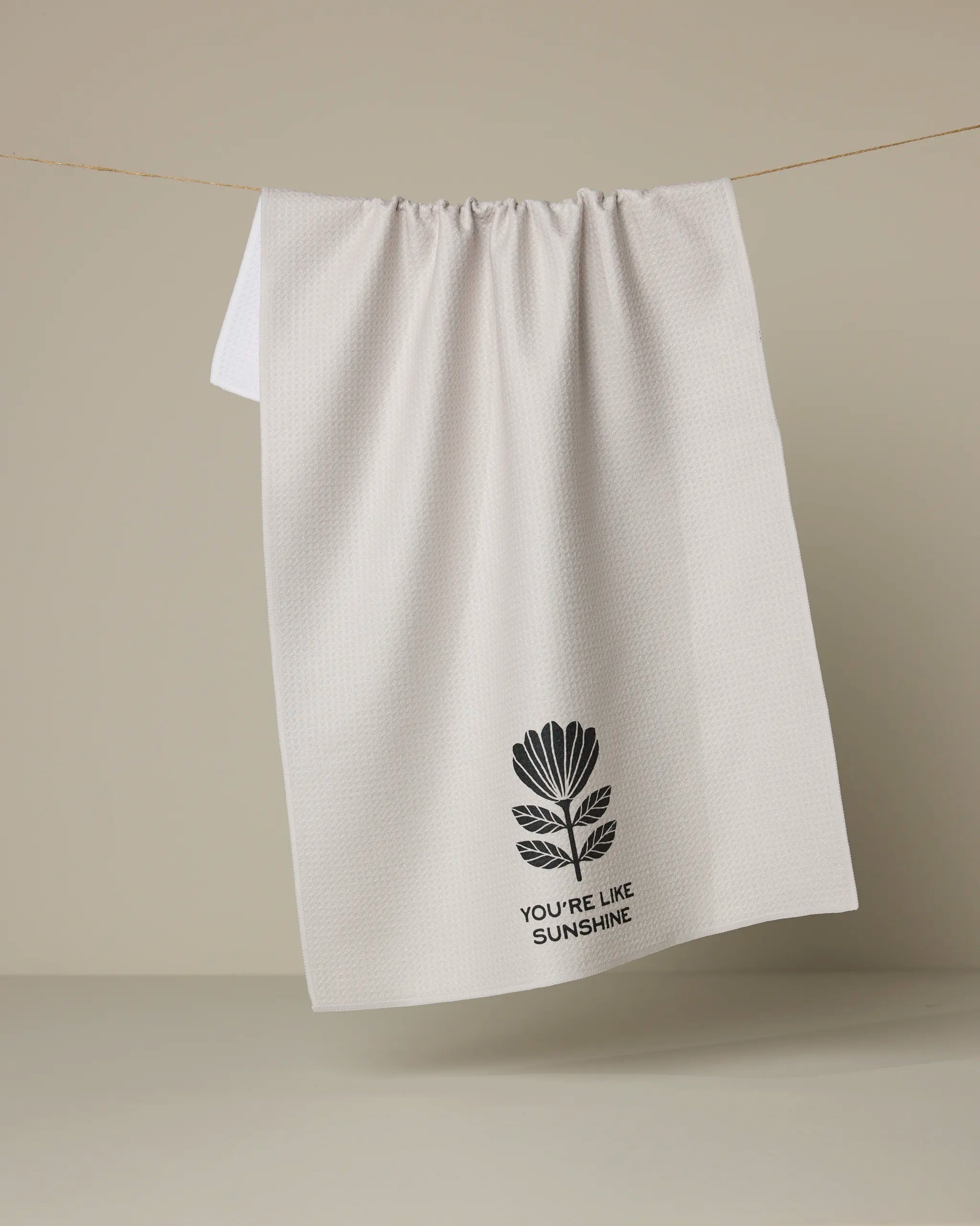 The Solveig white tea towel by Anna Christian, crafted from recycled materials, hangs on a line against a beige background. It displays a black flower graphic above the text "YOURE LIKE SUNSHINE.