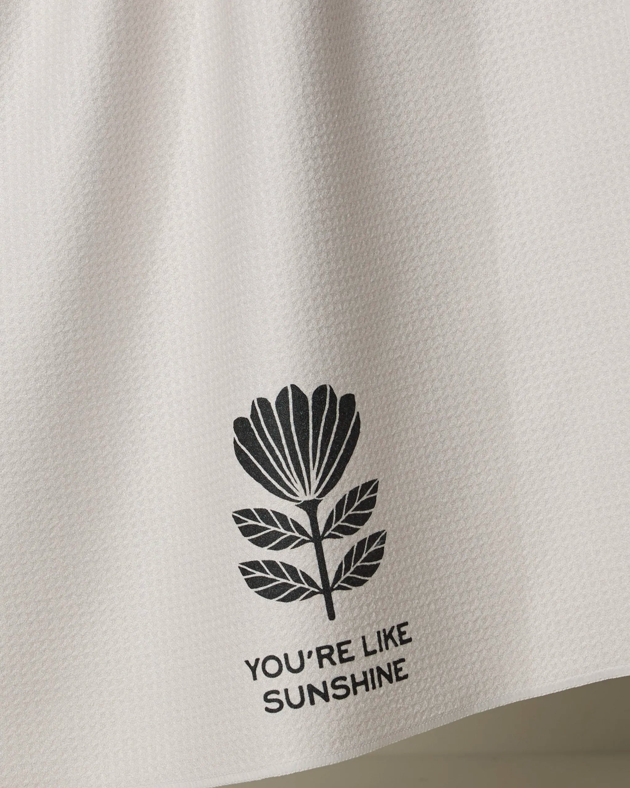 The Solveig tea towel by Anna Christian is white and textured, made from recycled materials, with a black floral illustration and “YOU’RE LIKE SUNSHINE” printed underneath.