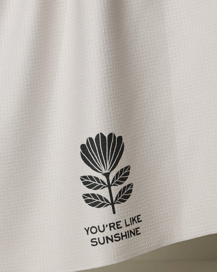 The Solveig tea towel by Anna Christian is white and textured, made from recycled materials, with a black floral illustration and “YOU’RE LIKE SUNSHINE” printed underneath.
