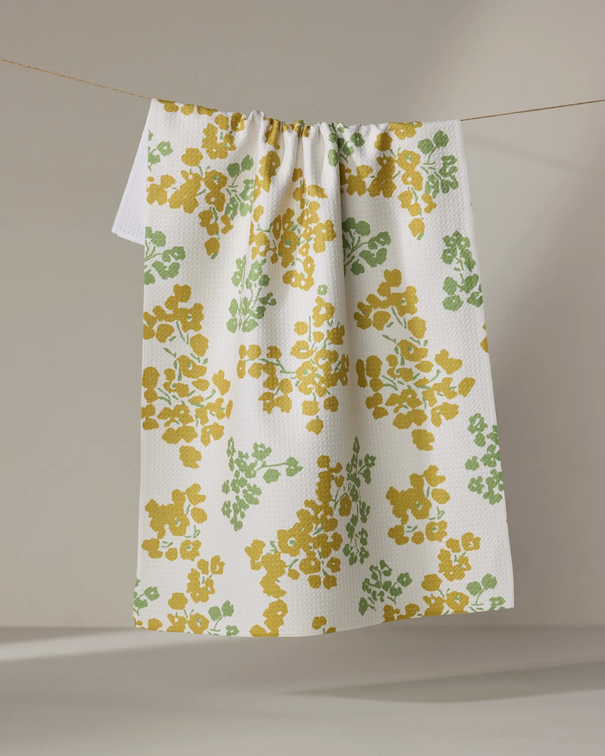 The Geometry Sonnet Butter tea towel, featuring a textured surface and yellow-green floral patterns, hangs draped over a clothesline against a neutral background.