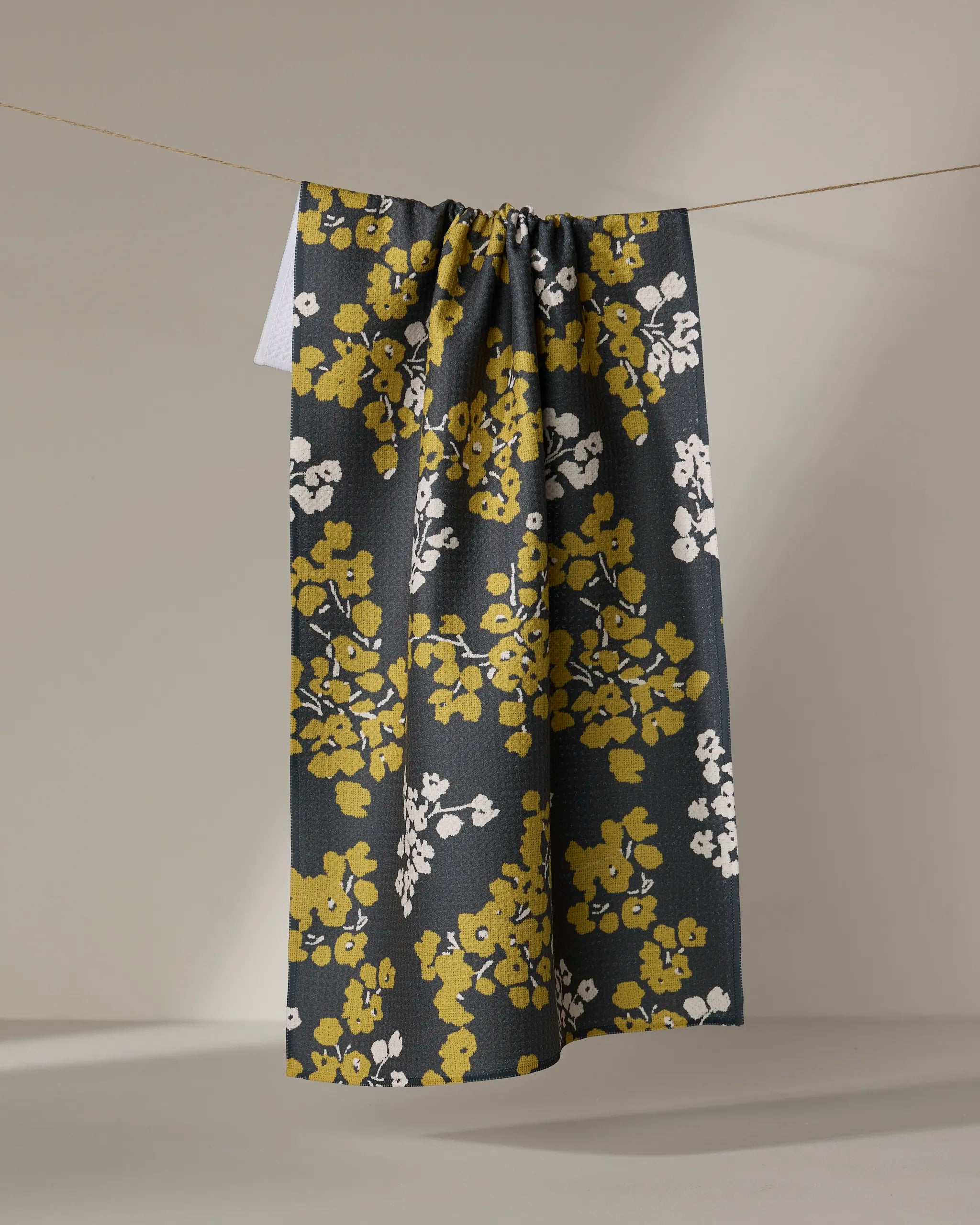 The Geometry Sonnet Ember tea towel, featuring a yellow and white floral pattern on a dark background and made from recycled materials, hangs against a plain, neutral backdrop.