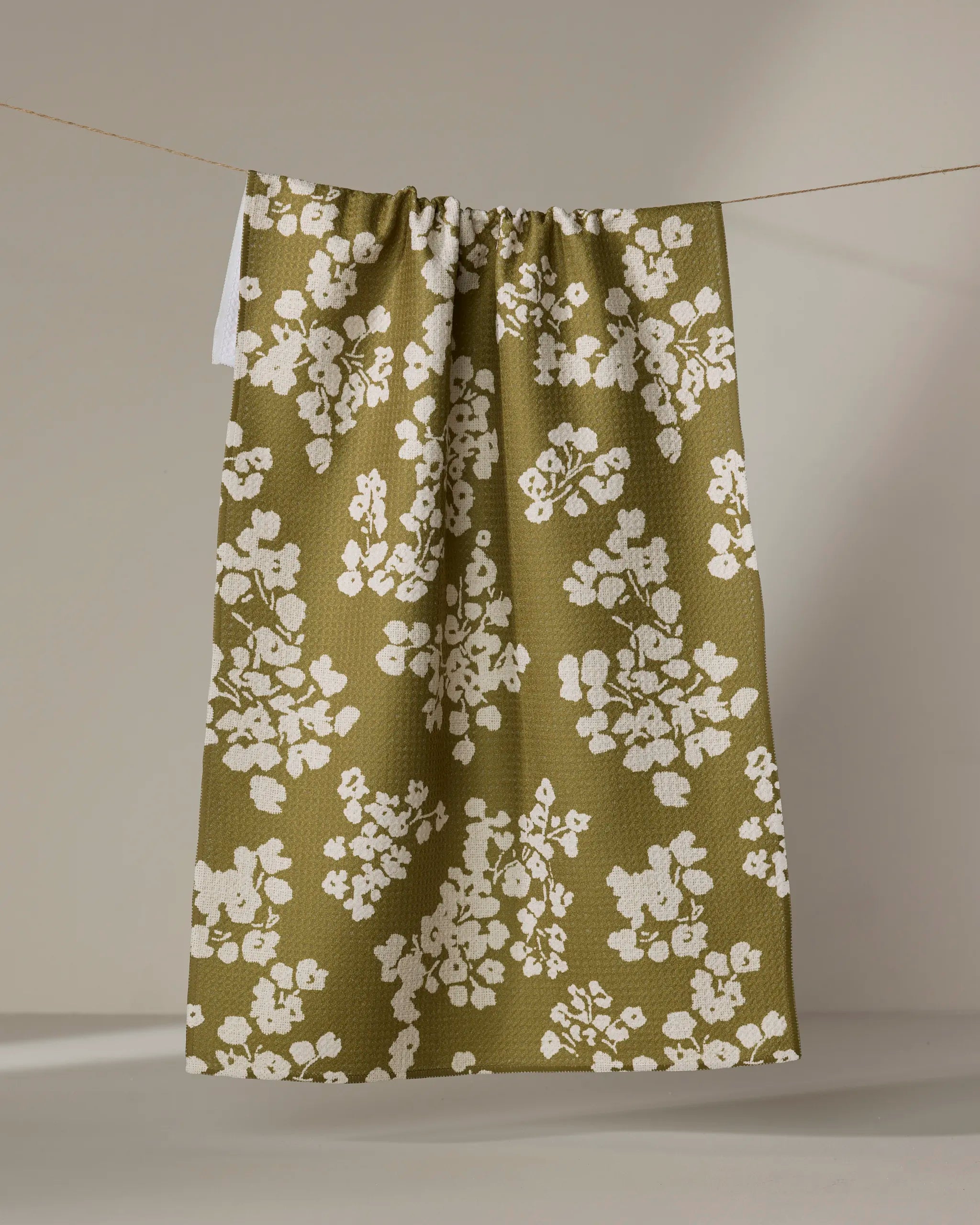 The Geometry Sonnet Olive tea towel, featuring a white floral pattern on an olive green background, hangs from a string against a neutral backdrop.