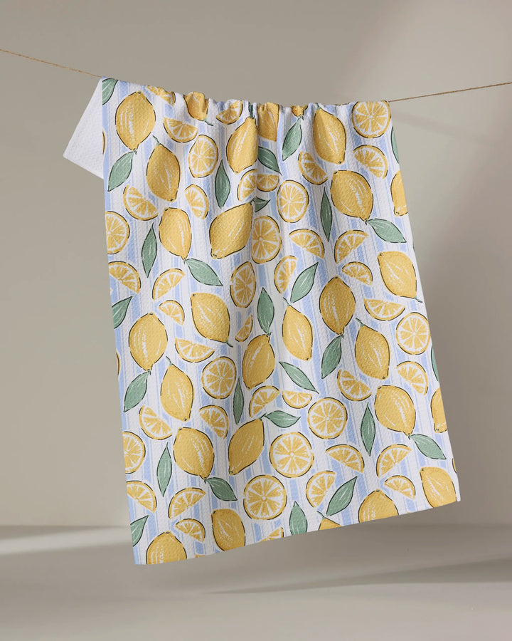 The Sour Lemon Stripes recycled kitchen towel by Hufton Studio features whole and sliced yellow lemons with green leaves, hanging from a line against a neutral background.
