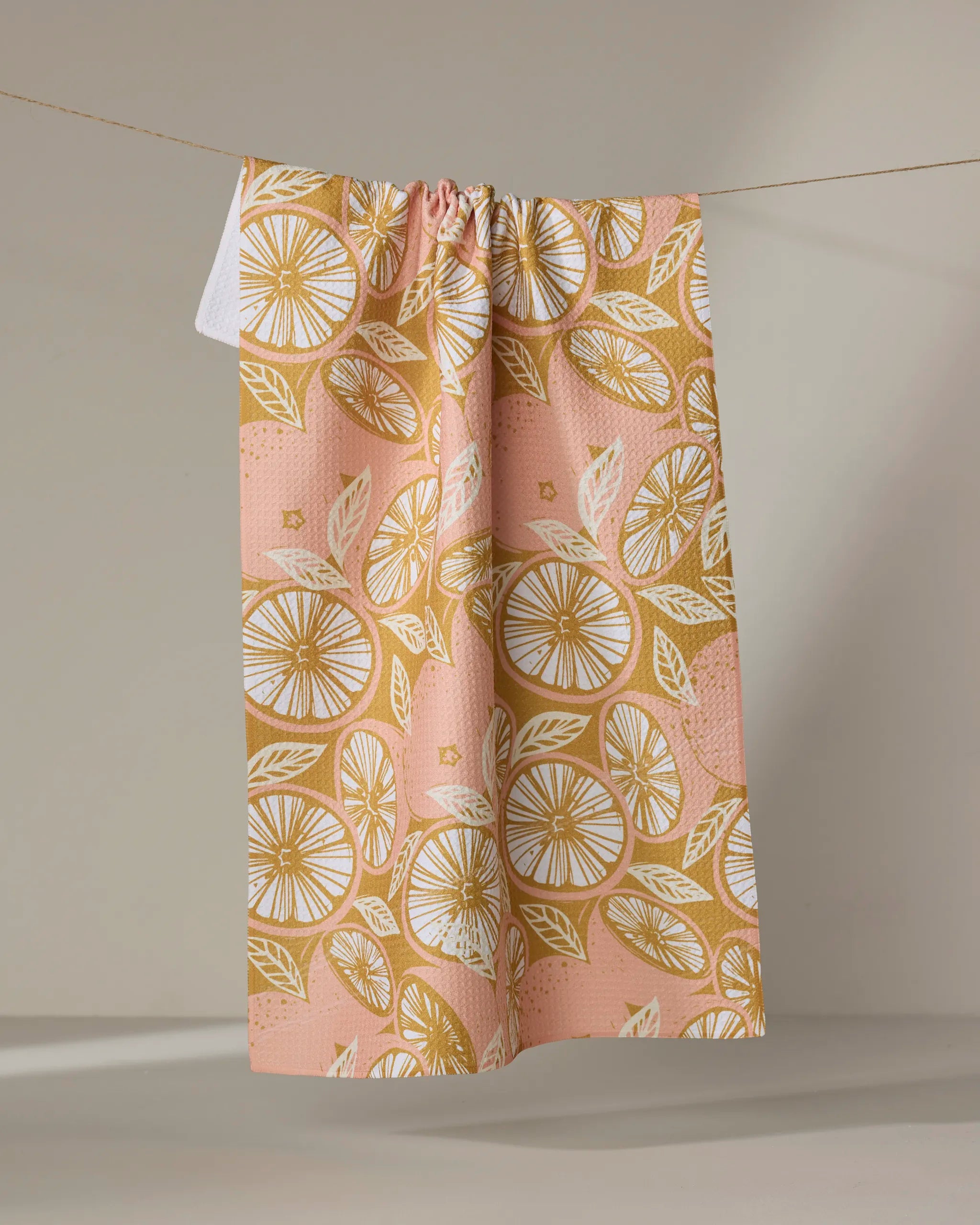 The Elisabeth Wing Spanish Grapefruit recycled kitchen towel, featuring a pink and mustard yellow citrus slice and leaf pattern, hangs draped over a clothesline against a neutral background.