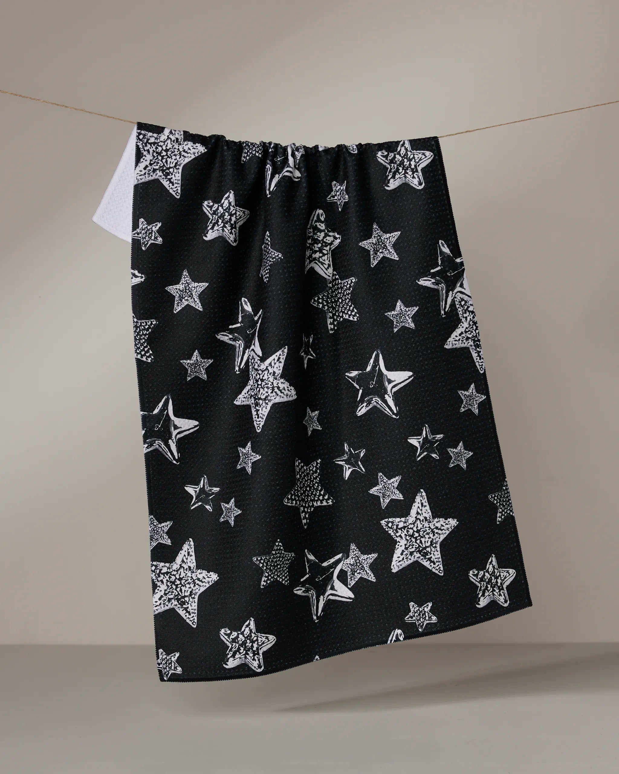 The Geometry Sparkling Night tea towel features a white star pattern on black and hangs from a clothesline against a neutral background—an eco-friendly kitchen towel ideal for everyday use.
