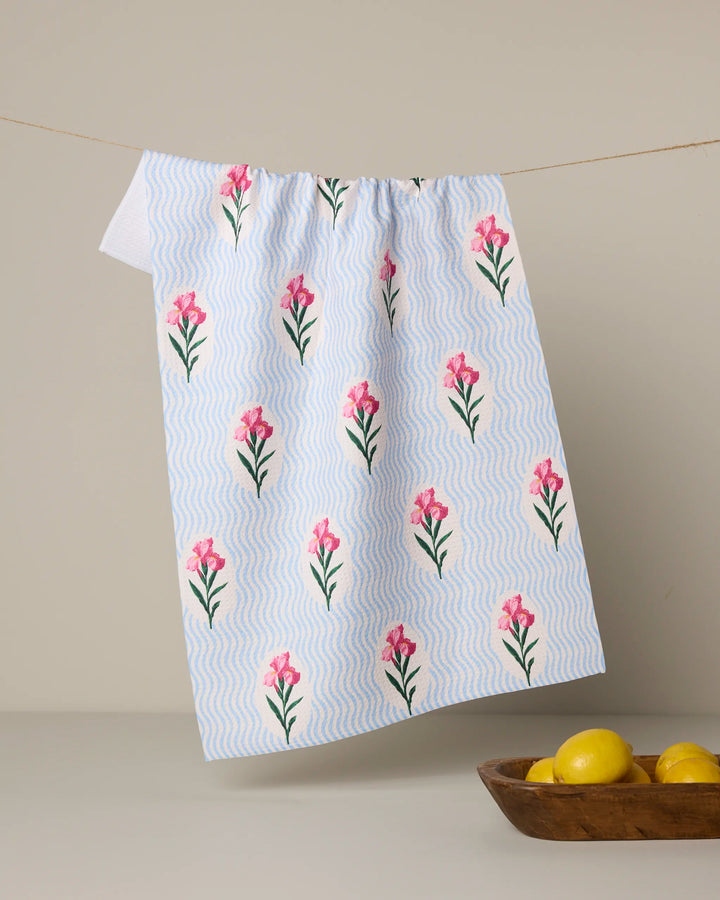 The Geometry "Spatial Bloom" tea towel, featuring a wavy blue and pink floral print on white recycled fabric, hangs on a line. Below, a wooden bowl with three yellow lemons rests against a beige backdrop.