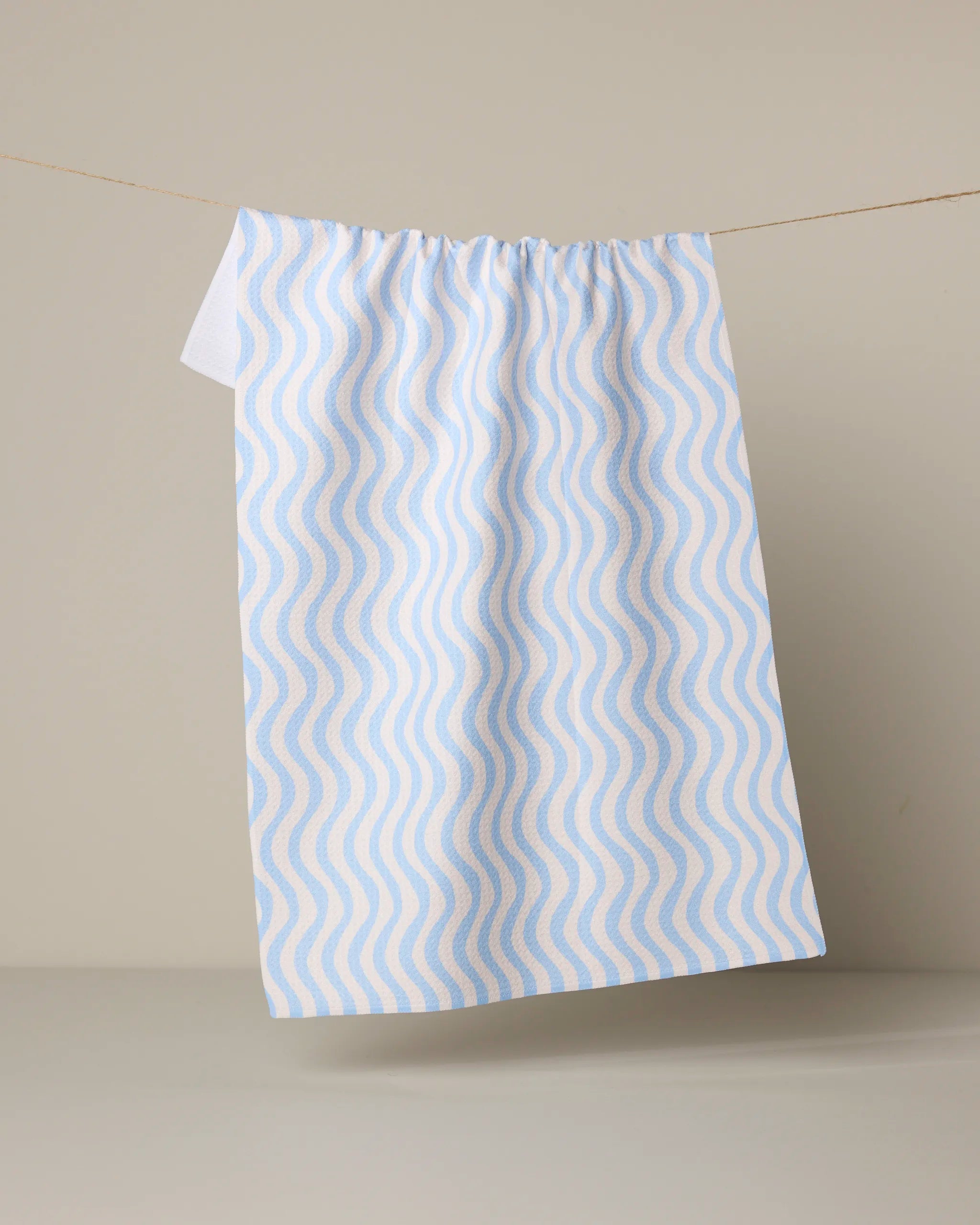 The Geometry Spatial tea towel, featuring a wavy blue and beige design and made from quick-drying recycled materials, hangs from a thin string against a plain light beige background.