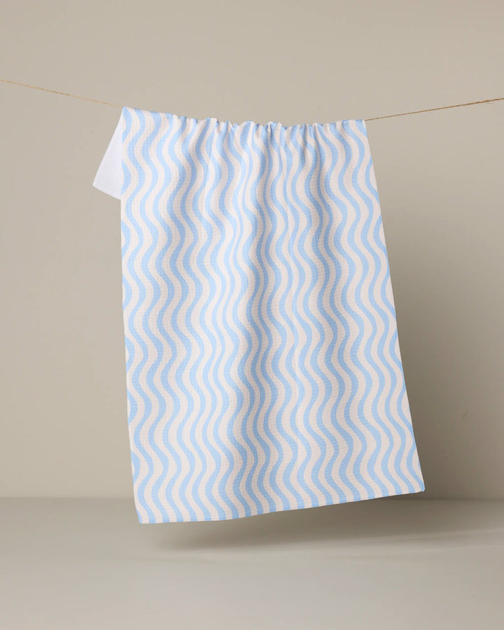 The Geometry Spatial tea towel, featuring a wavy blue and beige design and made from quick-drying recycled materials, hangs from a thin string against a plain light beige background.