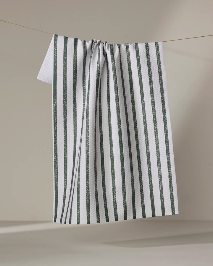A Becki Owens "Spearmint" tea towel made of white Geoweave fabric with vertical dark green stripes hangs draped on a string, forming gentle folds against a light beige background.
