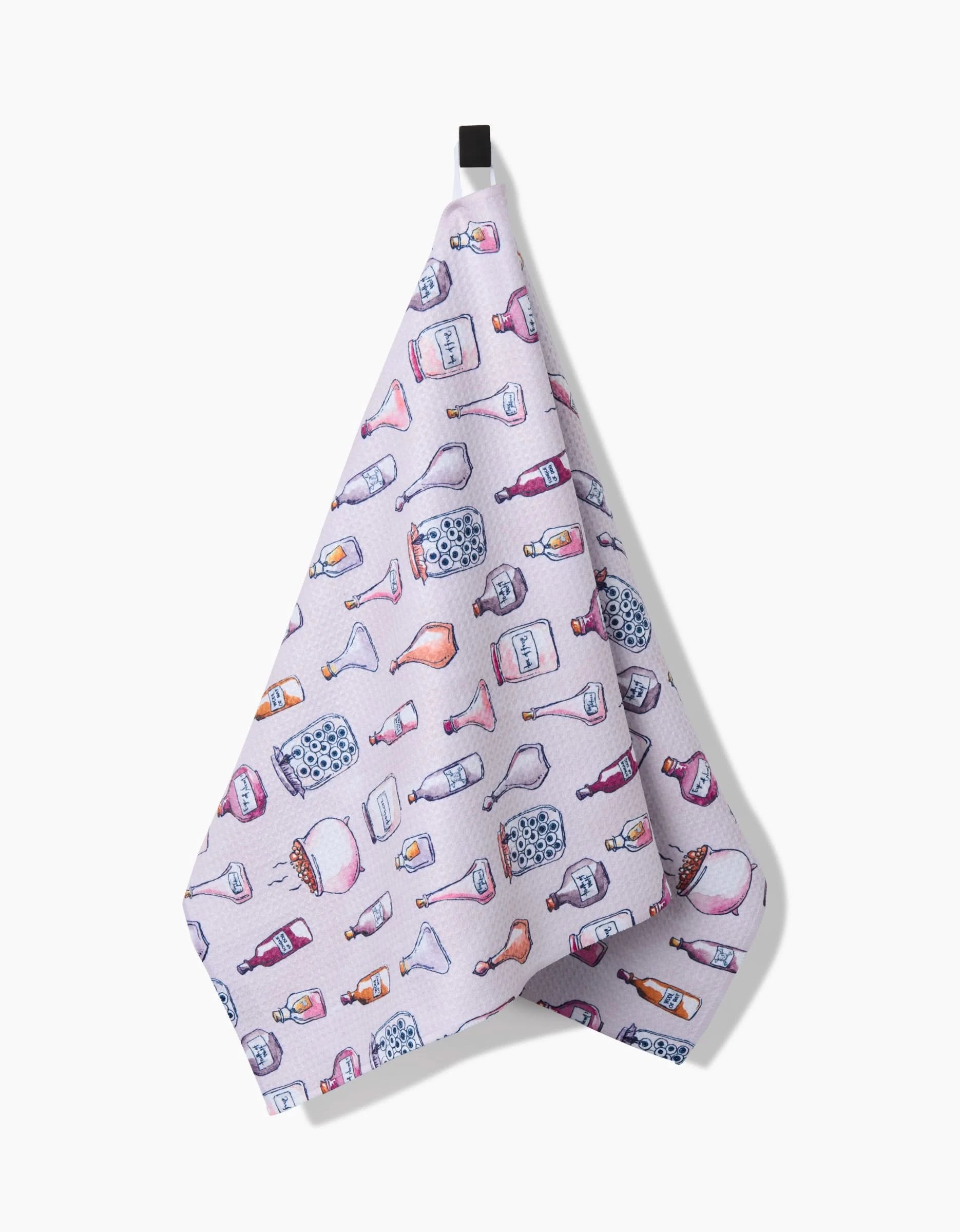 A pink recycled kitchen towel from Maayan Alper-Swan’s Spells Potions collection hangs from a black hook, showcasing illustrated bottle vases, jars, and containers in blue, purple, and orange.