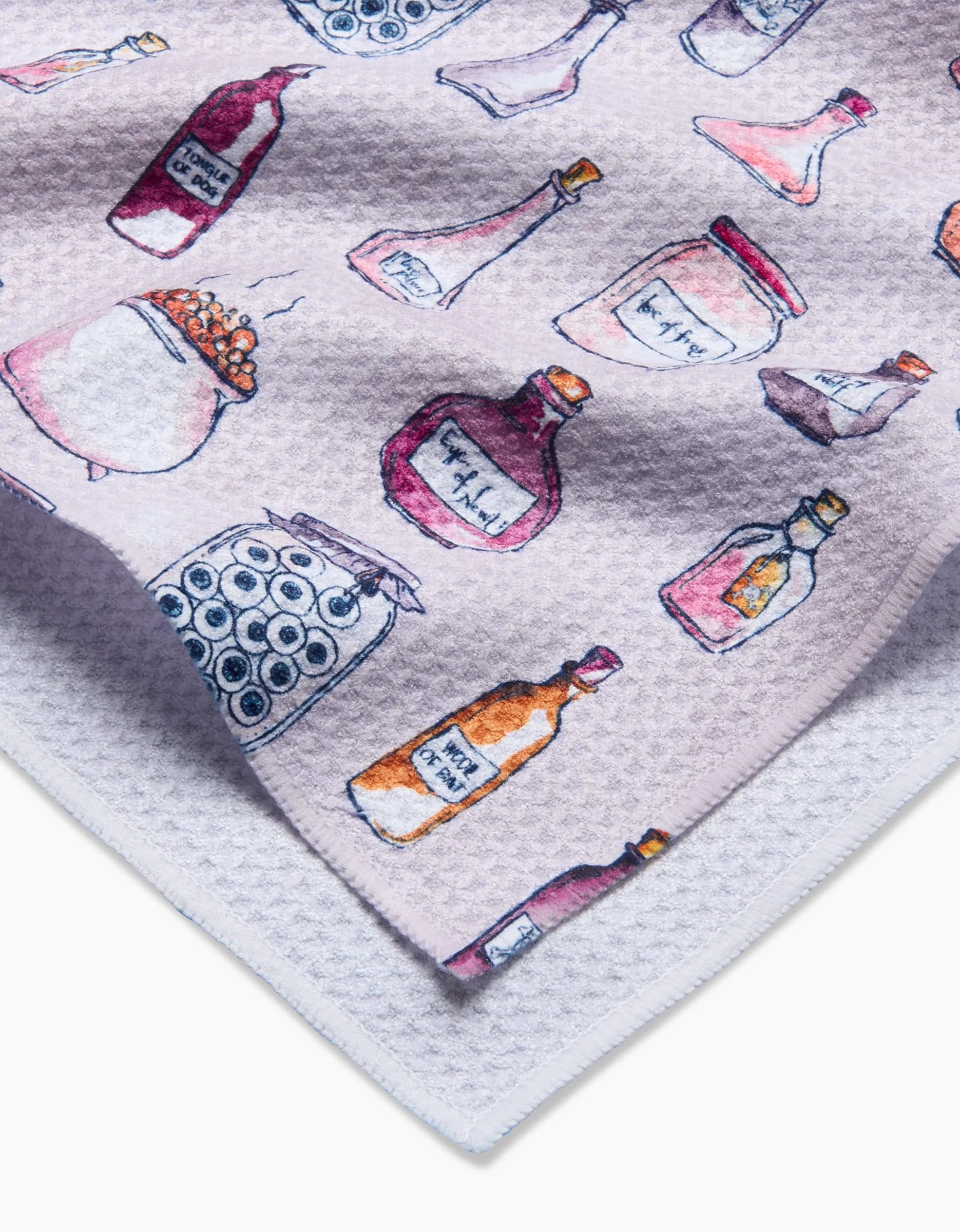 The Spells Potions kitchen towel by Maayan Alper-Swan is white, textured, and recycled, featuring a colorful hand-painted pattern of assorted potion bottles and jars—some labeled—scattered randomly across the surface.