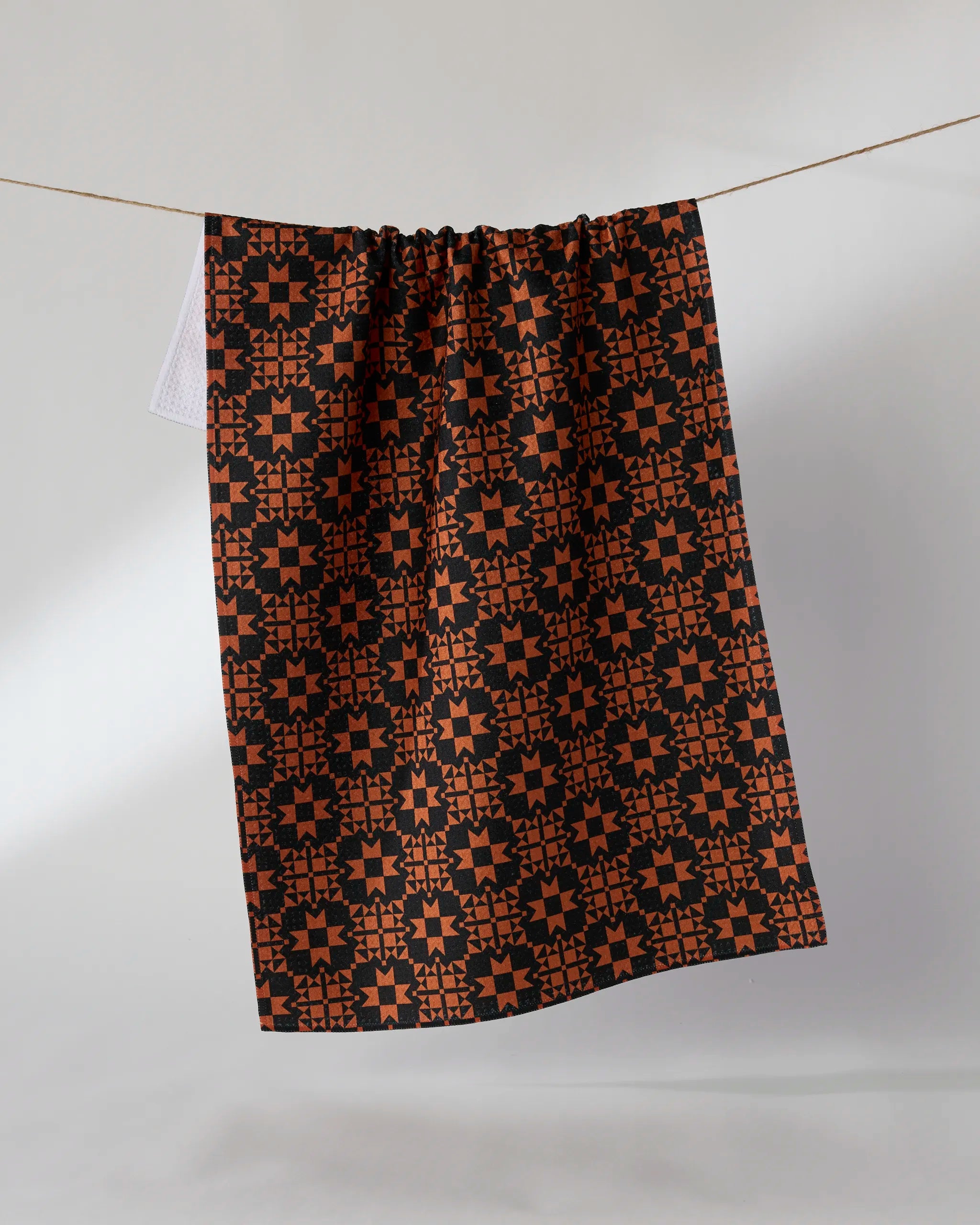 The Spellwork tea towel by Geometry, featuring a bold orange and black geometric star pattern, hangs on a clothesline against a light background, highlighting its stylish look and absorbent kitchen towel qualities.