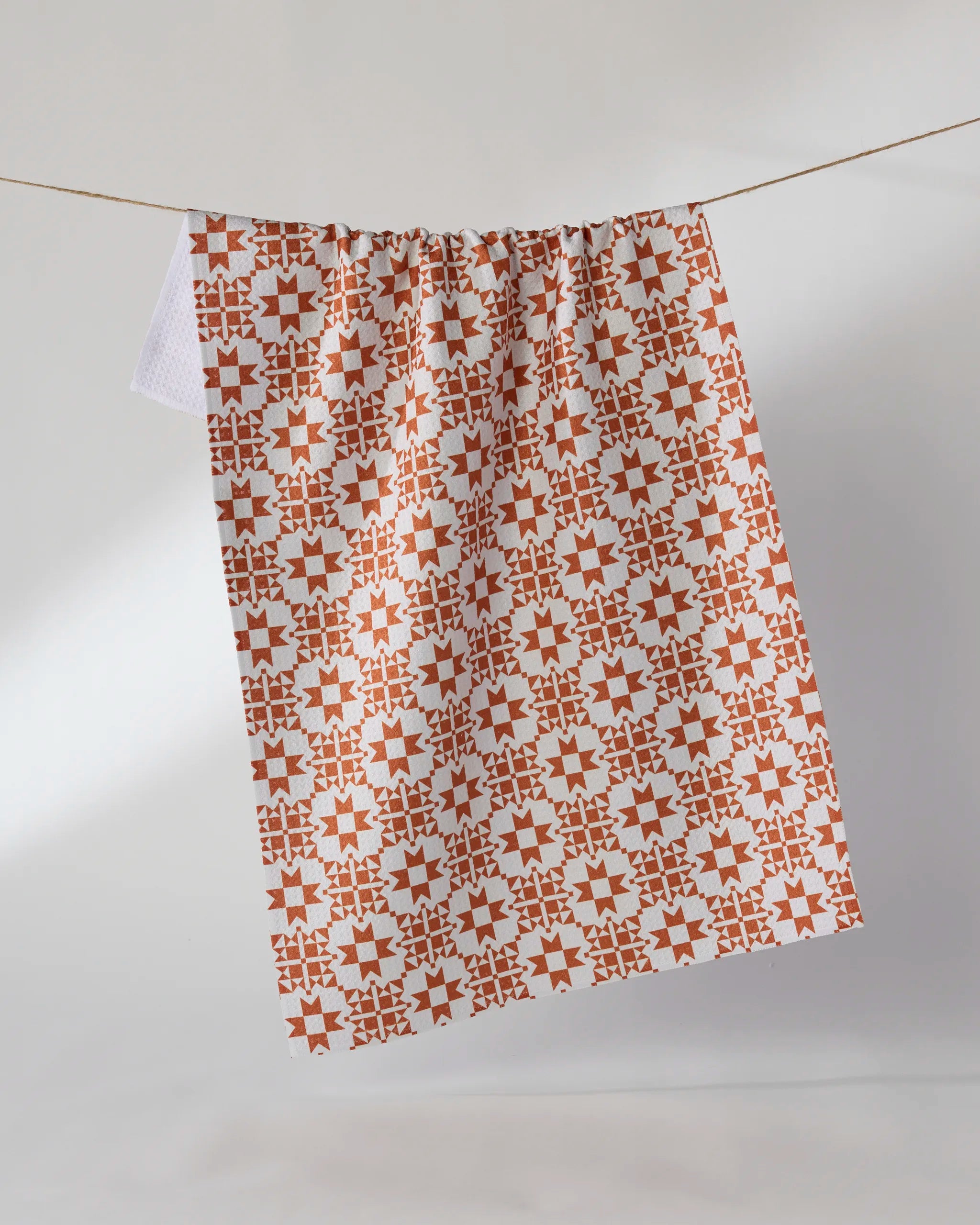 The Geometry Spellwork recycled kitchen towel, featuring a red and white geometric pattern, hangs on a clothesline against a plain white background.