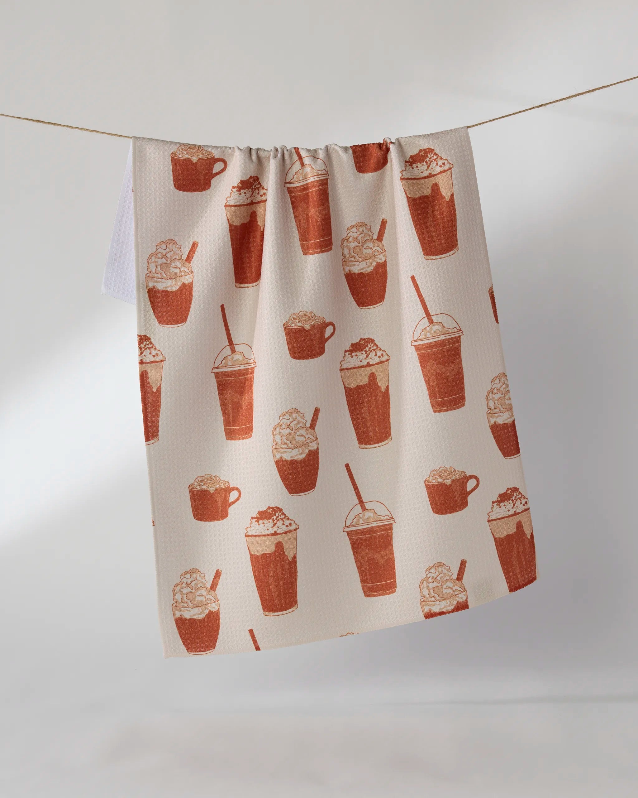 The Geometry Spice Season tea towel features iced coffee, frappuccino, and hot chocolate drinks in brown hues on beige. Ultra-absorbent and stylish, it’s perfect for your kitchen and shown hanging against a light background.