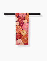 The Spicy Pumpkins tea towel by Rebecca Bobko, featuring vibrant orange, red, and pink flowers, hangs on a black bar against a white background. Made from quick-drying recycled materials.