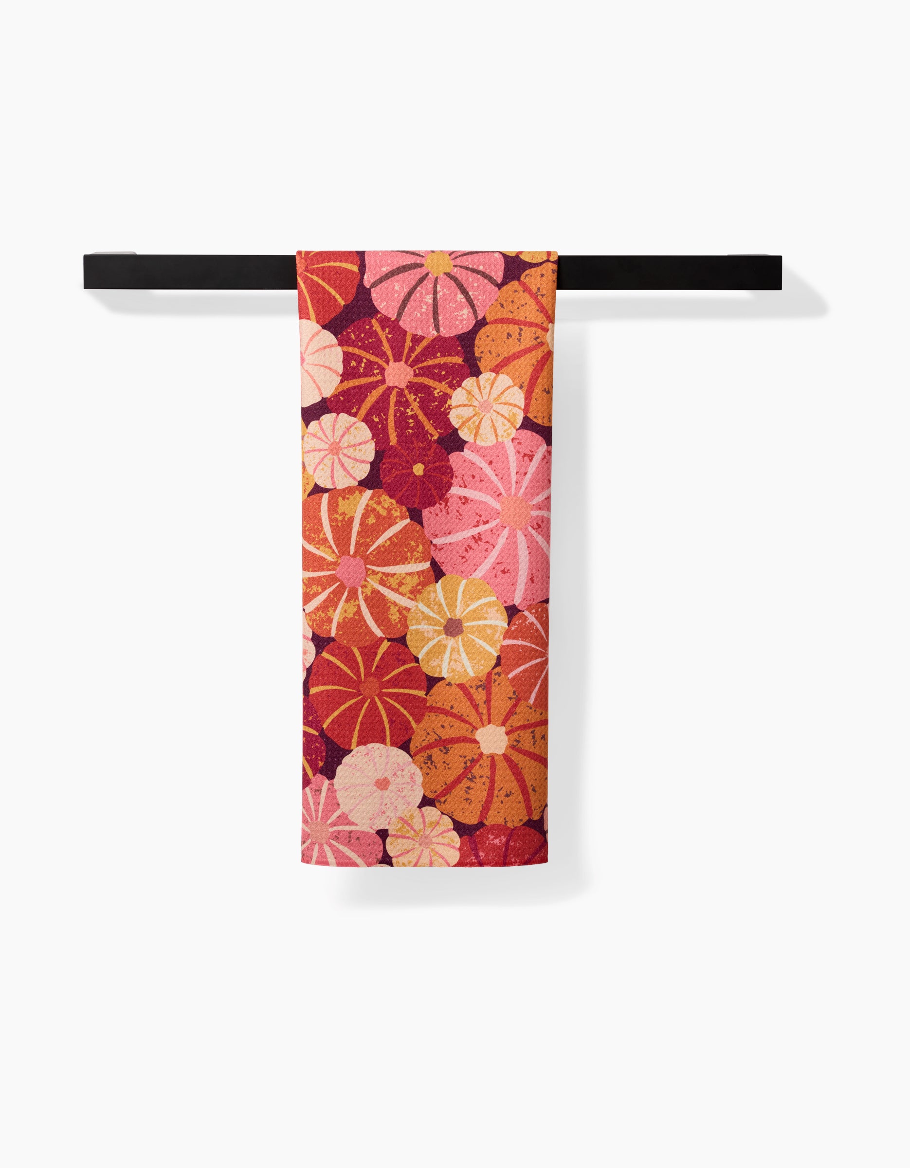 The Spicy Pumpkins tea towel by Rebecca Bobko, featuring vibrant orange, red, and pink flowers, hangs on a black bar against a white background. Made from quick-drying recycled materials.