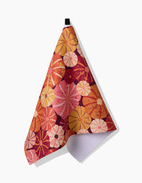 The Spicy Pumpkins tea towel by Rebecca Bobko features an orange, pink, and red floral pattern. Hanging from a black loop against a white background, it’s made from quick-drying recycled materials with a white textured underside.