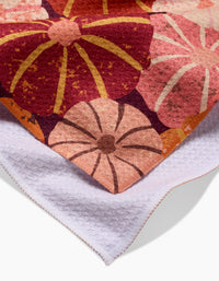 The Spicy Pumpkins tea towel by Rebecca Bobko features a vibrant floral pattern in red, pink, and orange on one side and plain white textured recycled fabric on the other.