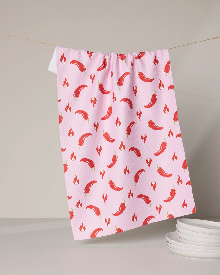 The Geometry Spicy tea towel, made from recycled materials, features a quick-drying pink fabric with red chili peppers and flame icons, hanging on a clothesline above a stack of white plates.