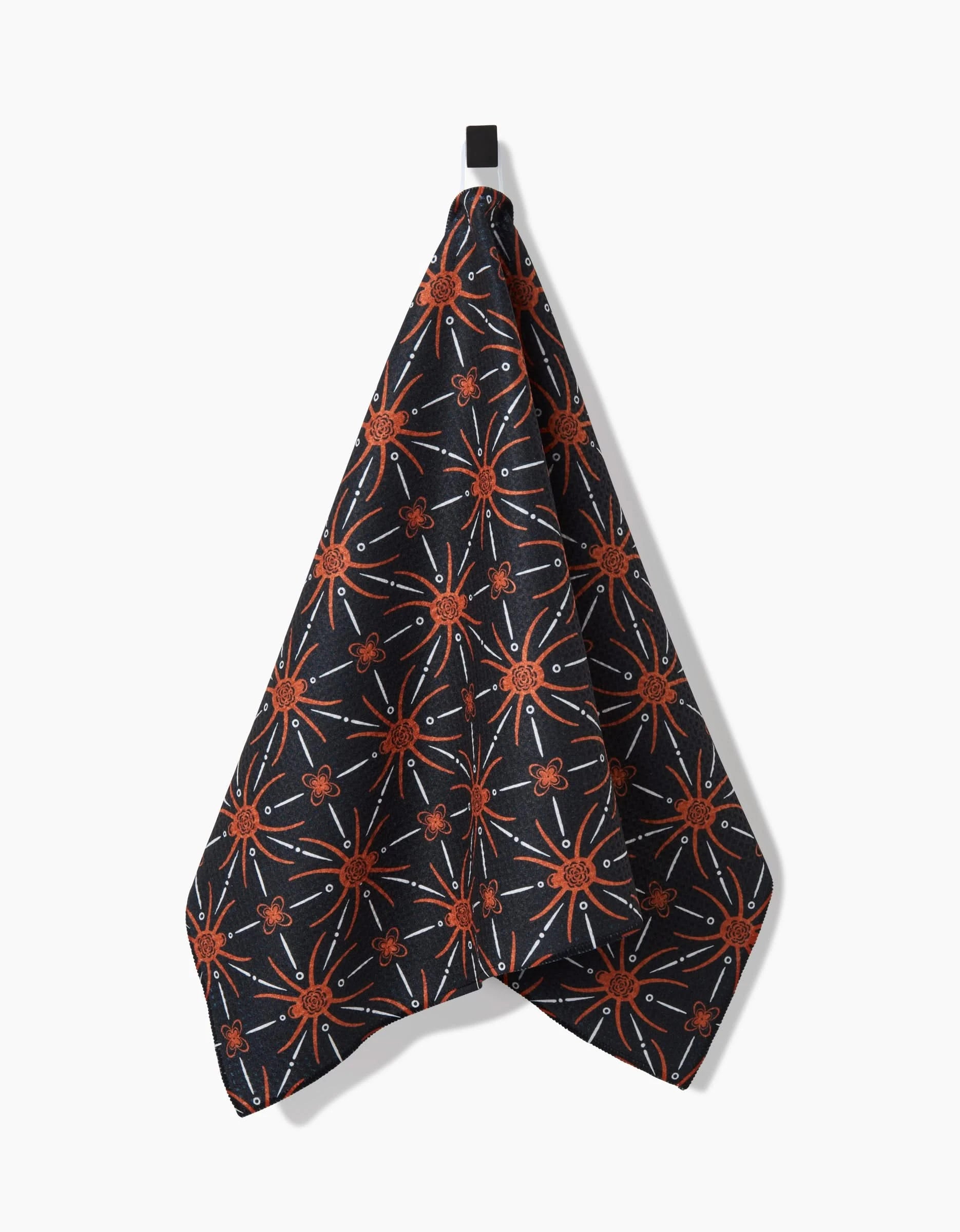 The Sarah Daugherty Spider Geometric towel, featuring a dark base with orange starburst and clover shapes, hangs from a black loop against a plain white background.