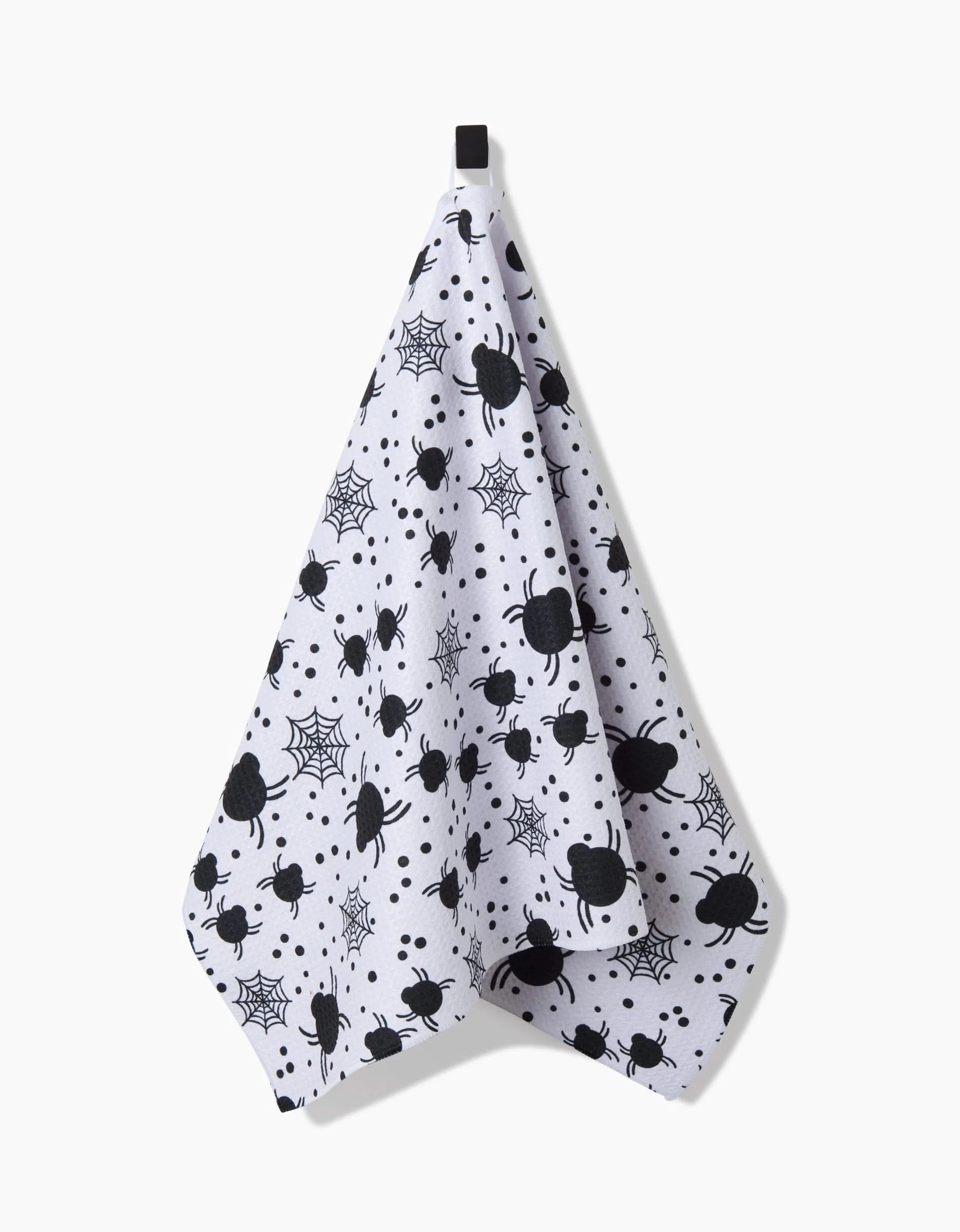 The Spiders For Days tea towel by Charlie Rowan Designs features black spider prints and webs on white, hanging from a black loop. Crafted from recycled materials, this ultra-absorbent towel is stylish and eco-friendly.