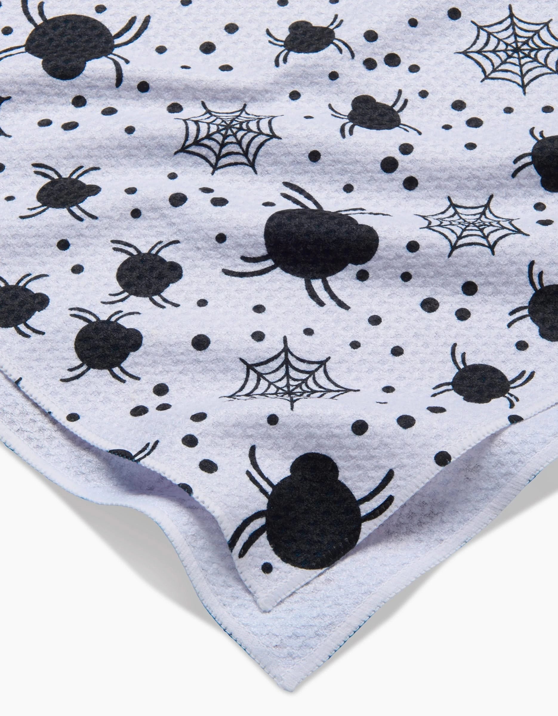 A close-up of Charlie Rowan Designs’ “Spiders For Days” white kitchen tea towel, showcasing black spiders, webs, and dots with a folded corner of its ultra-absorbent fabric.