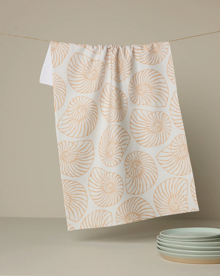 The Geometry Spira recycled kitchen towel, featuring a peach nautilus shell pattern, hangs on a line. In front, a stack of light gray plates rests on a beige surface with a neutral background.