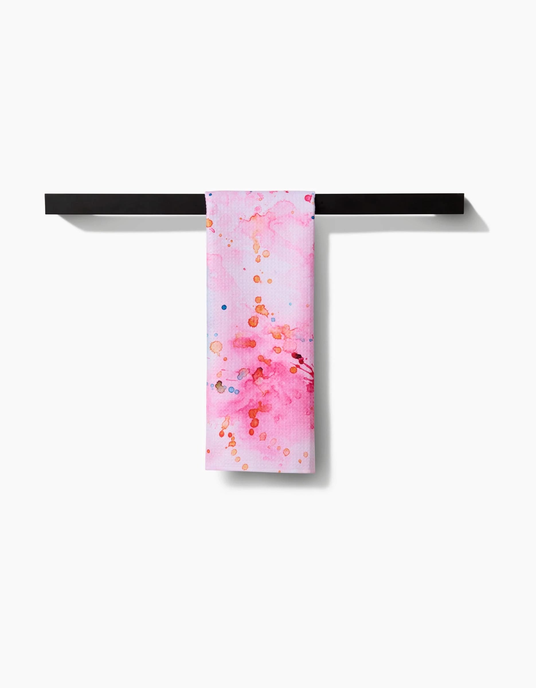 The Splash of Love towel by Sara Mari, featuring pink and orange watercolor splashes on white ultra-absorbent fabric, hangs neatly on a black towel bar against a plain white background.