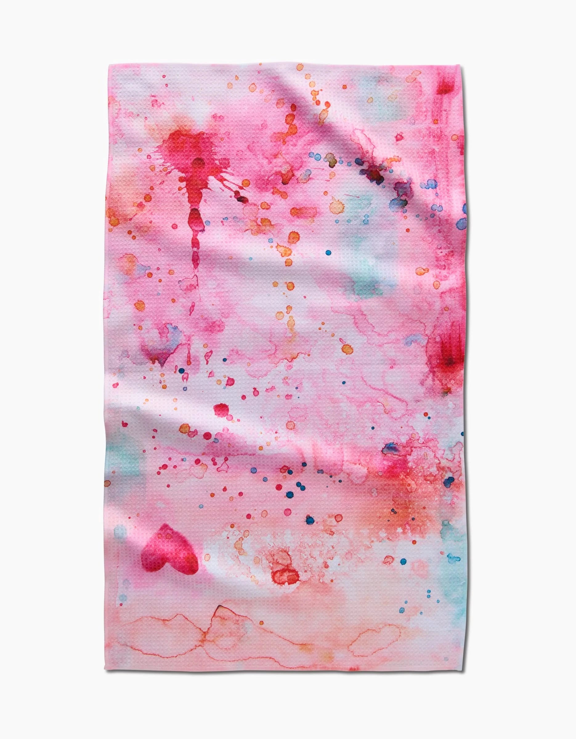 The Sara Mari Splash of Love kitchen towel is rectangular, white, and recycled, featuring abstract pink, red, blue, and orange splashes with a faint heart on the lower left. The edges have a slightly wrinkled finish.
