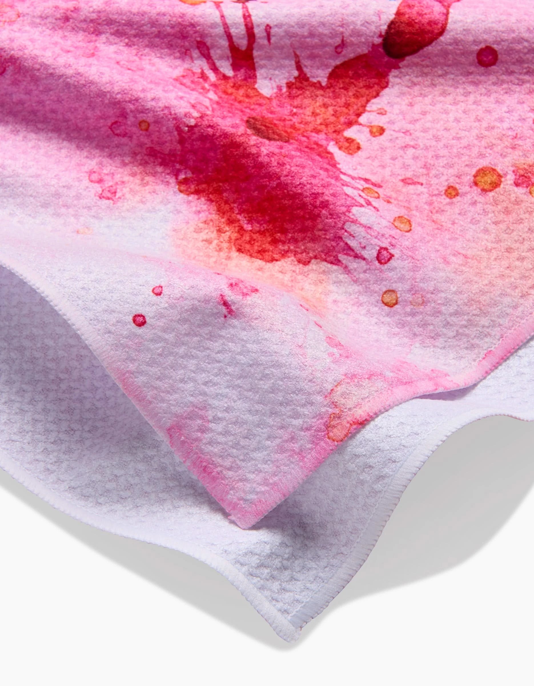 A close-up of Sara Maris Splash of Love towel, featuring ultra-absorbent textured white fabric with vibrant pink and red watercolor-style splashes. Slightly folded edges reveal the underside.