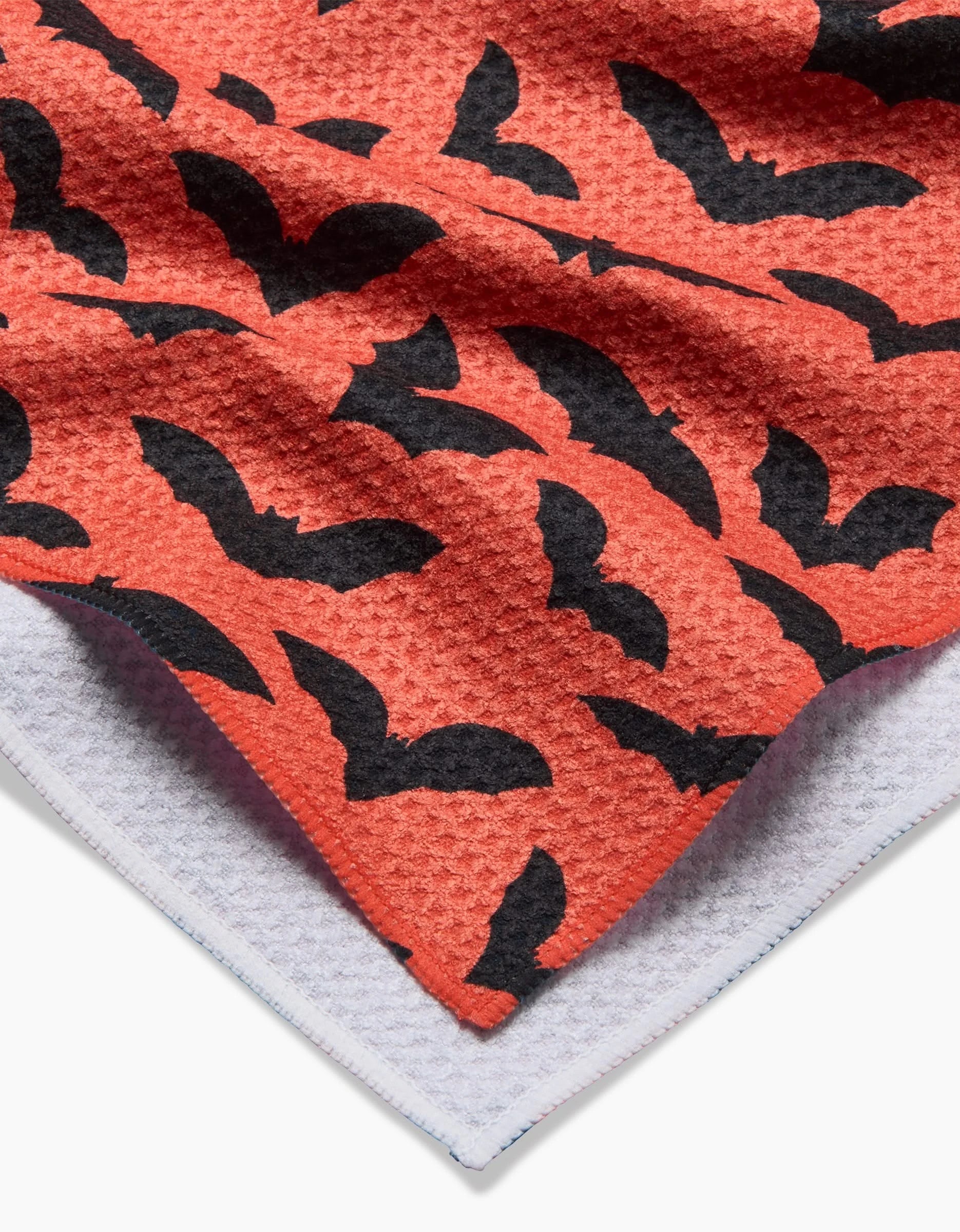 A close-up of the Rebecca Bobko Spooky Bats tea towel, featuring an orange fabric with a black bat pattern layered over a white towel. The quick-drying texture is clearly showcased.