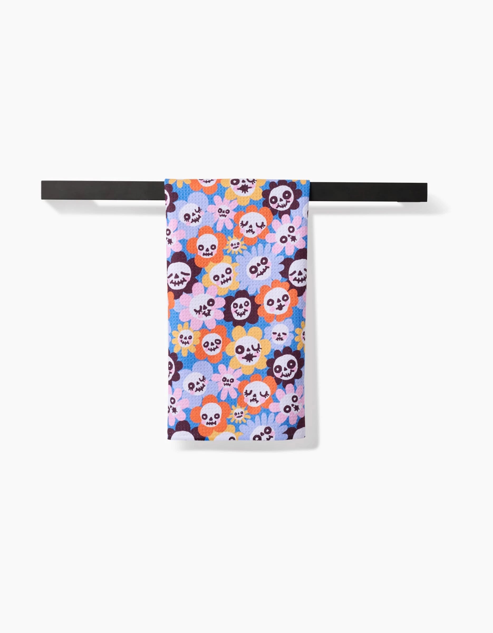 The Spooky Bouquet tea towel by So Lazo, featuring a vibrant floral and skull pattern in pink, orange, blue, and purple, hangs neatly on a black towel bar against a white background.