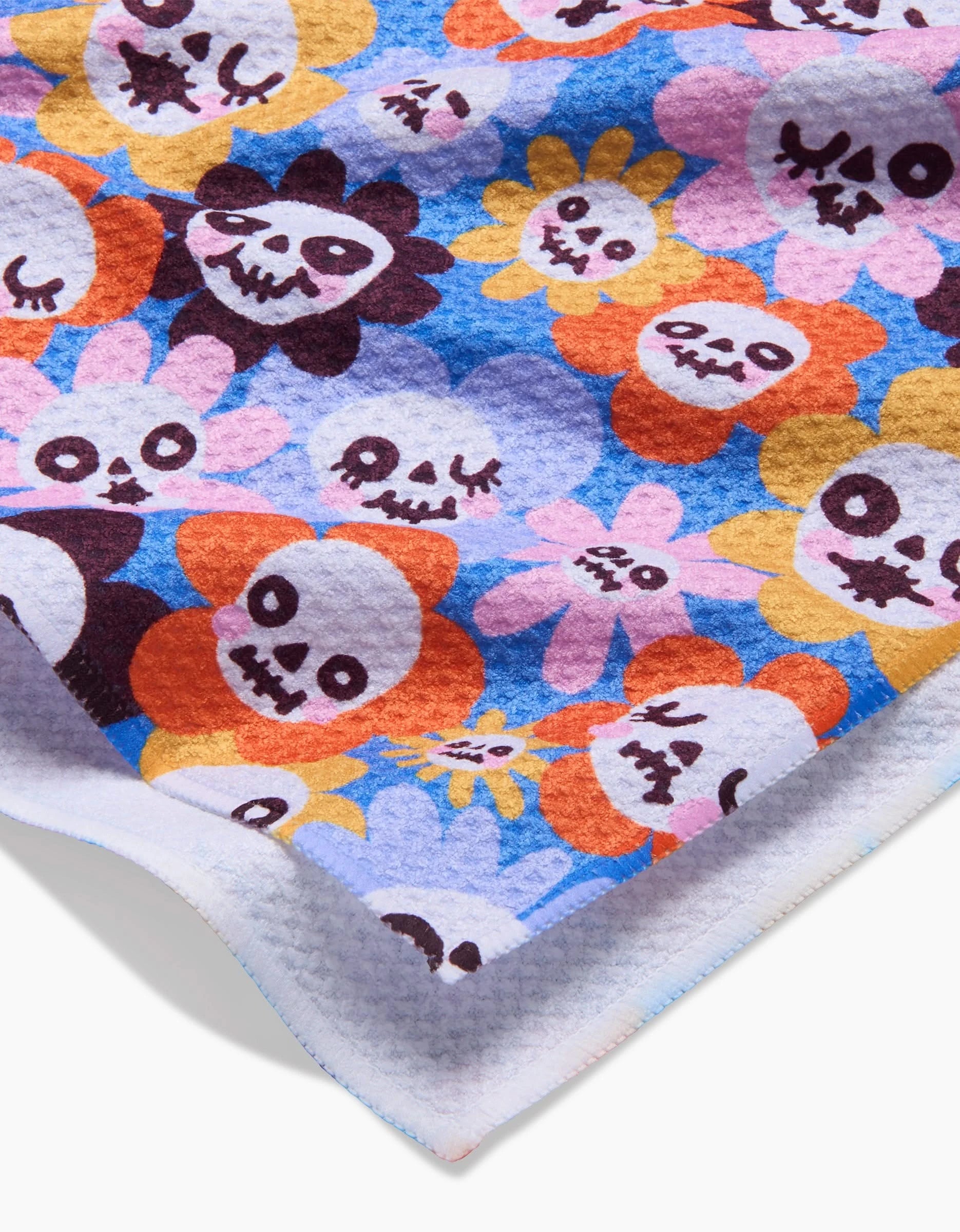 A close-up of So Lazo’s Spooky Bouquet towel shows its textured, quick-drying fabric and colorful cartoon skulls with flower petals in orange, yellow, purple, and pink on a blue background.