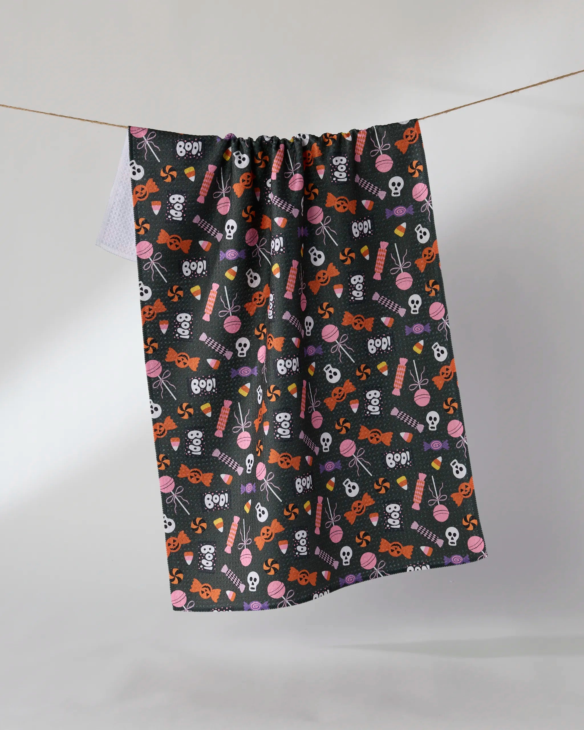 The Geometry Spooky Sweets tea towel, featuring a black quick-drying fabric with white skulls, orange jack-o’-lanterns, purple candy, bones, eyeballs, and pink bows in a Halloween pattern, hangs on a clothesline.