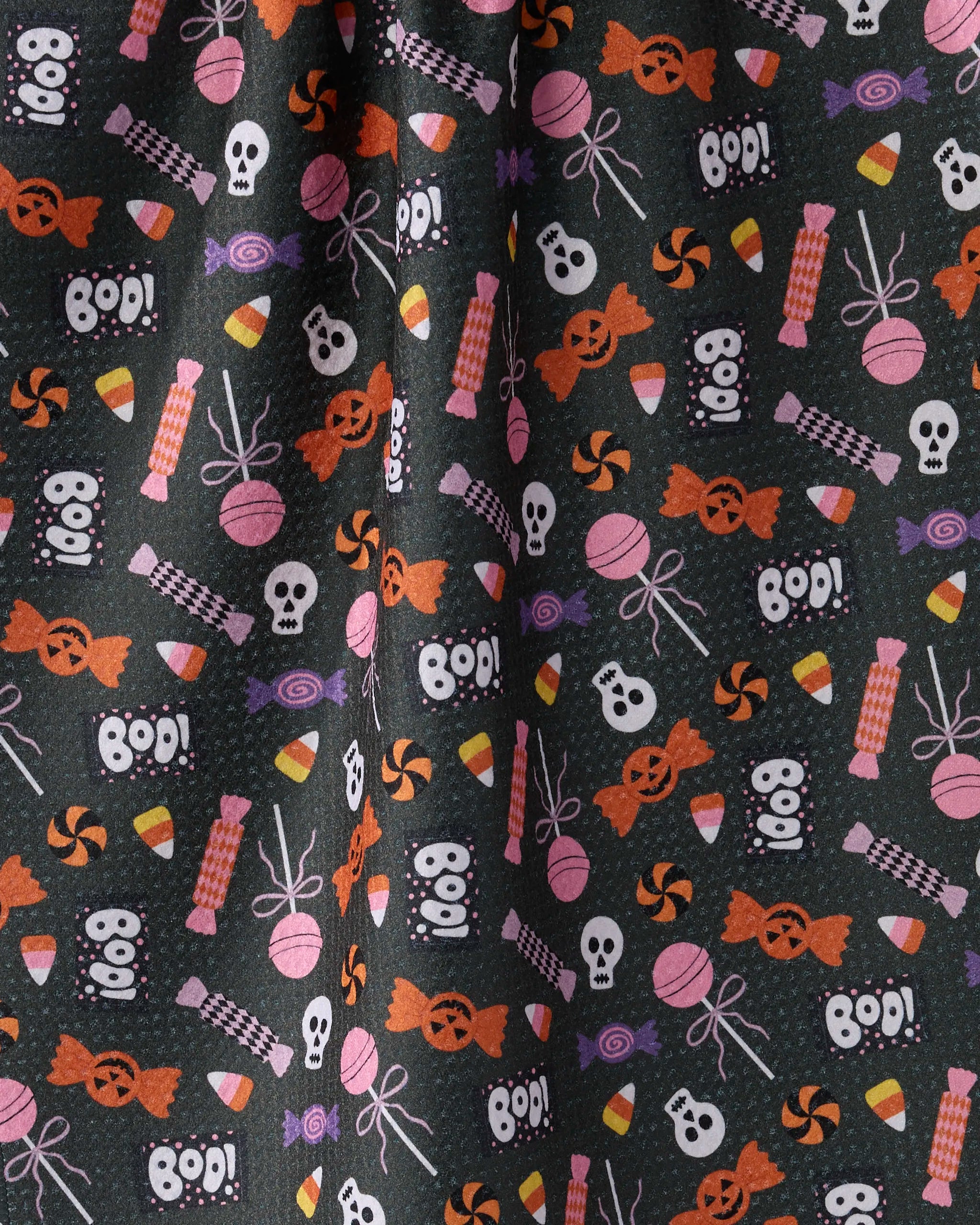The Geometry Spooky Sweets tea towel is quick-drying and features a Halloween pattern with skulls, “BOO!”, candies, and lollipops in orange, purple, white, and pink on a black background.