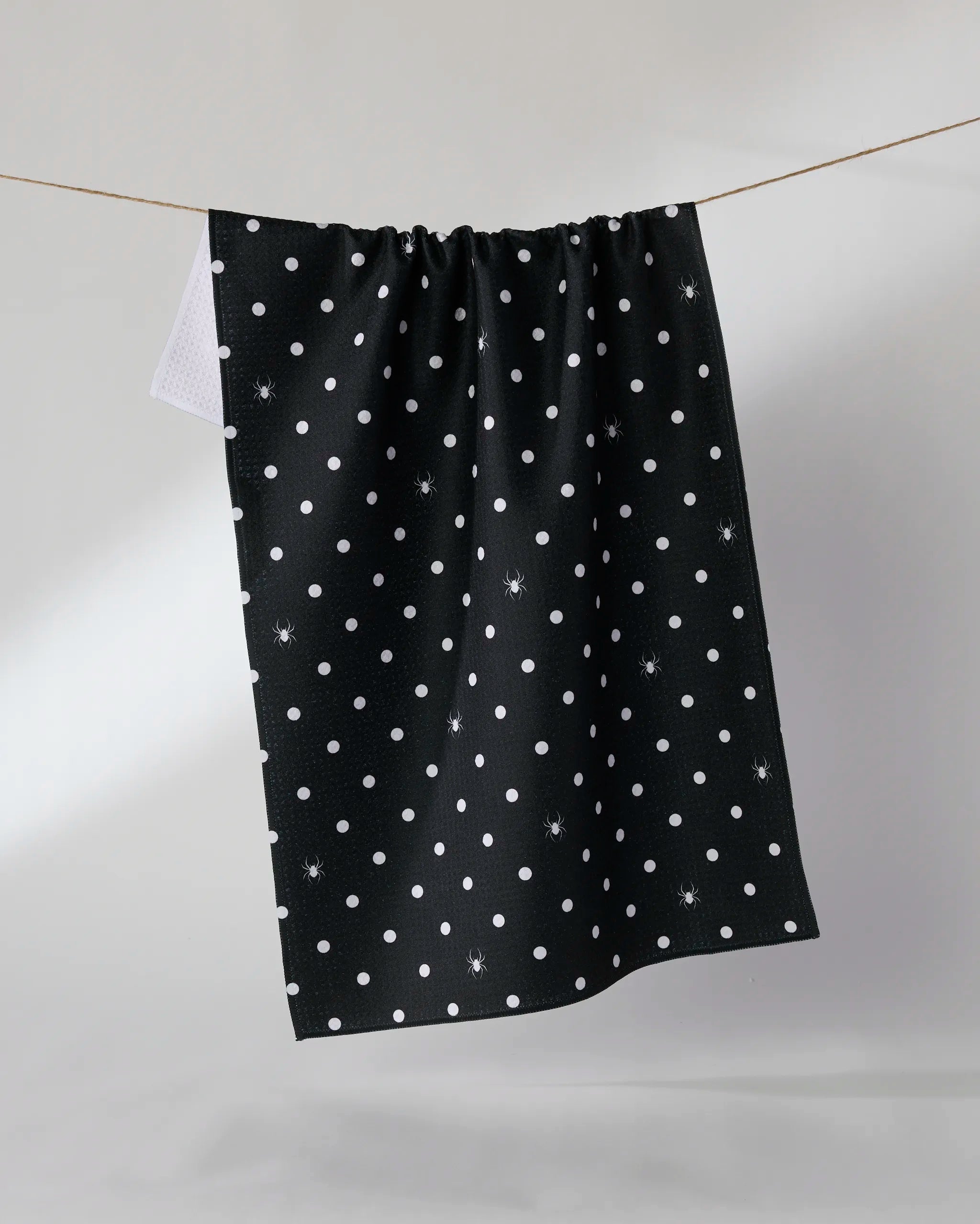 The Geometry Spookydots tea towel, featuring white polka dots on black and made from recycled materials, hangs on a clothesline against a plain light background.