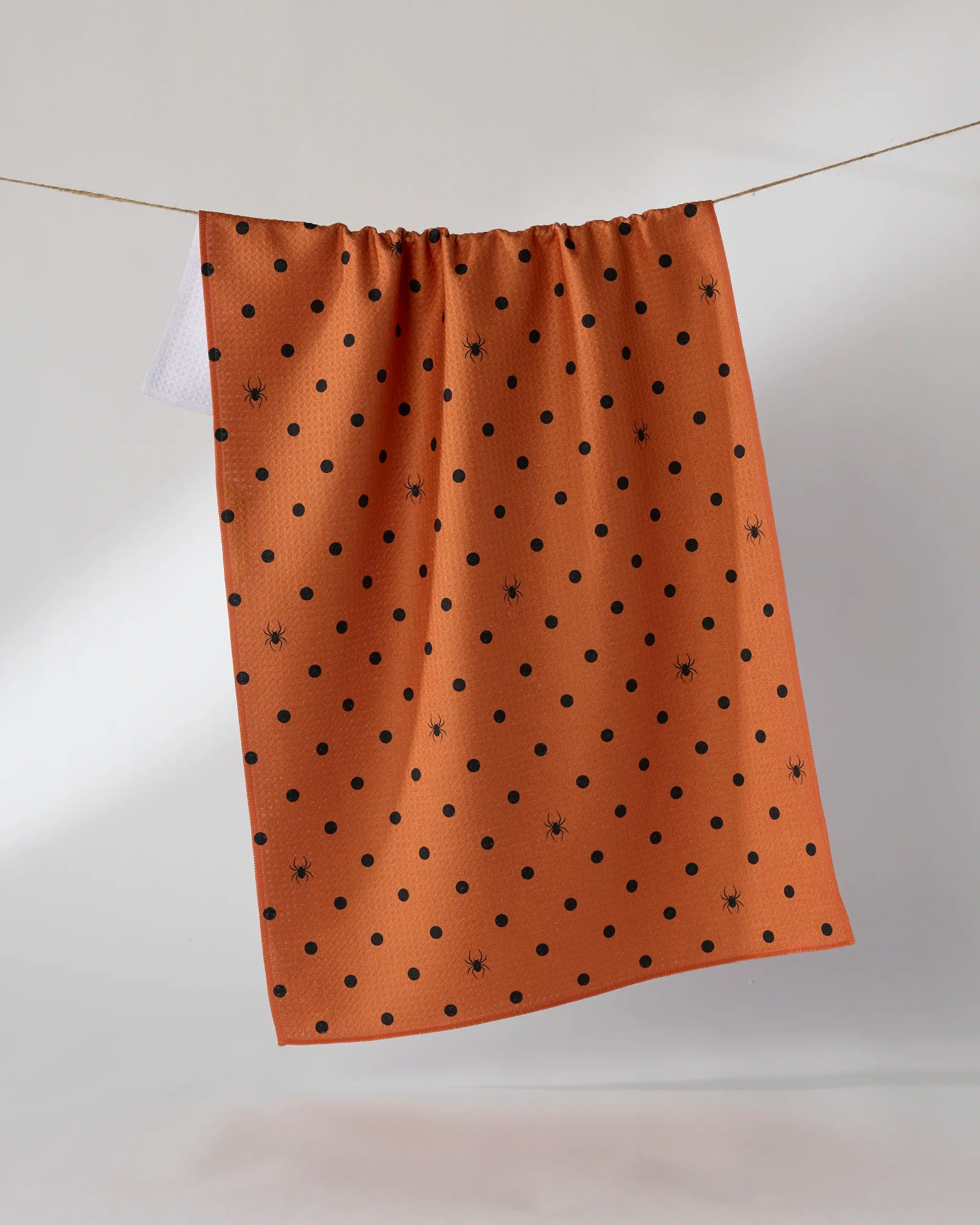 The Geometry Spookydots kitchen towel features black polka dots and small spider designs on an orange background, made from recycled materials, shown hanging on a clothesline against a plain light background.