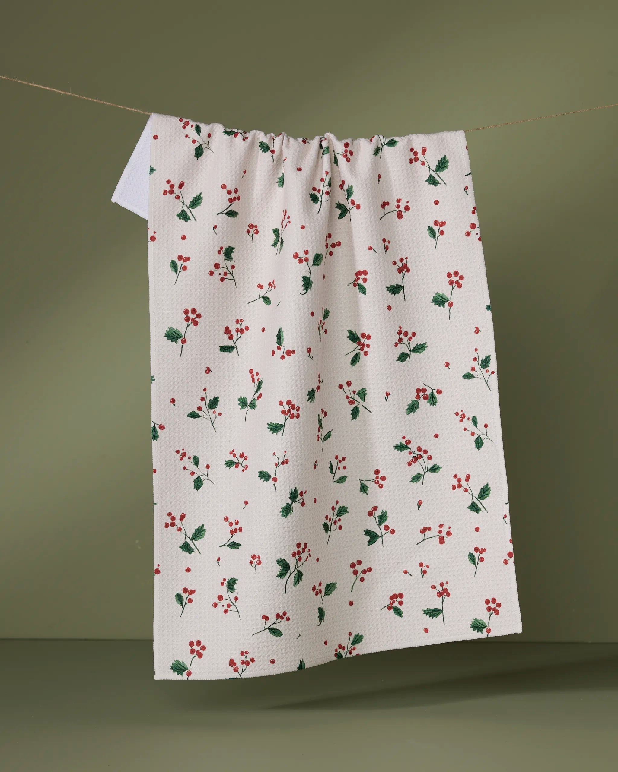The Geometry "Sprig" recycled white tea towel, featuring small red berries and green leaves, hangs from a string against a muted green background.