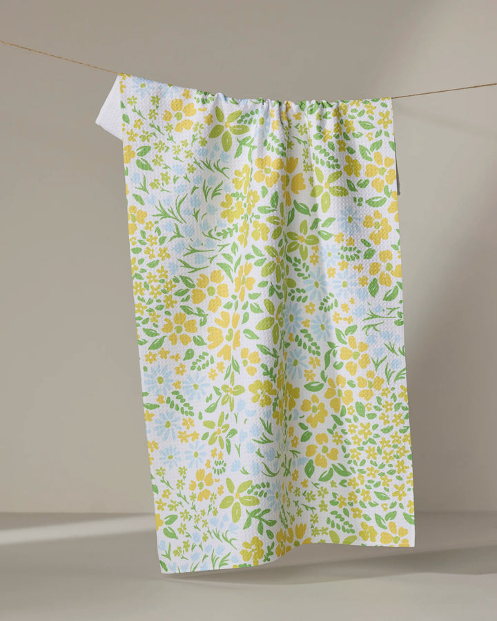 The Dabney Lee "Spring Awakening" tea towel, featuring a yellow and green floral pattern and made from ultra-absorbent recycled materials, hangs on a beige background.