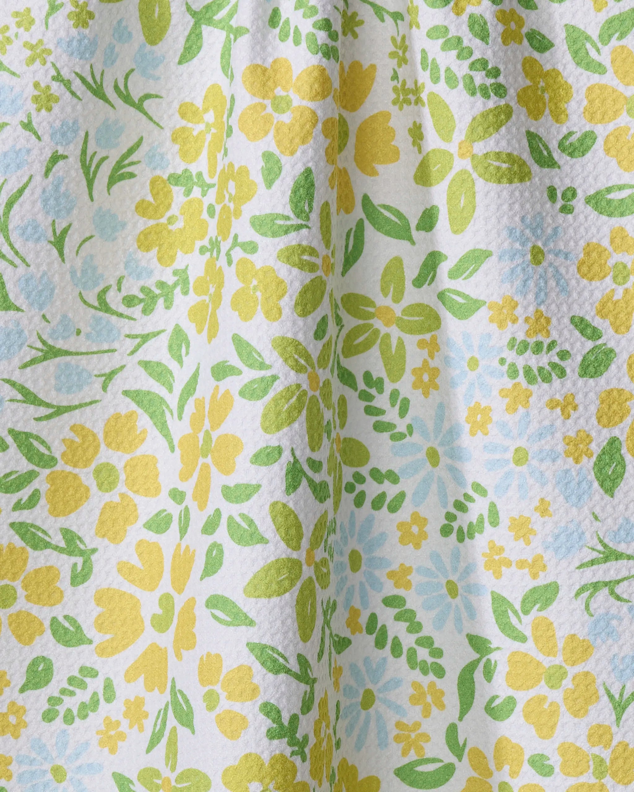 A close-up of Dabney Lee's "Spring Awakening" tea towel, ultra-absorbent with a textured surface, showcases a floral pattern in yellow, green, and blue, featuring various flowers and leaves with natural folds and shadows.