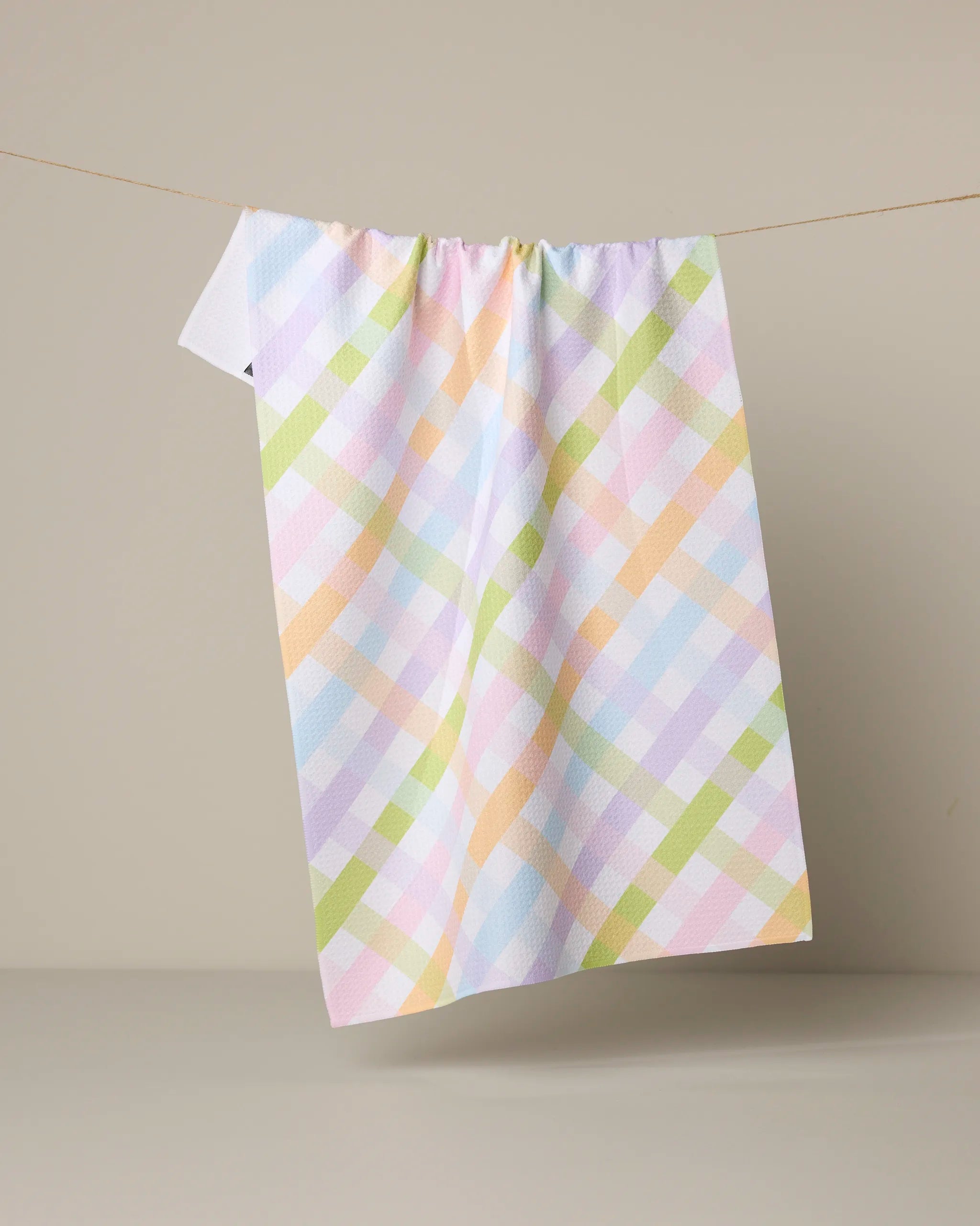 The Geometry Spring Basket tea towel, made from recycled materials, hangs on a clothesline against a beige background and features pastel plaid with intersecting pink, blue, green, orange, and yellow lines.