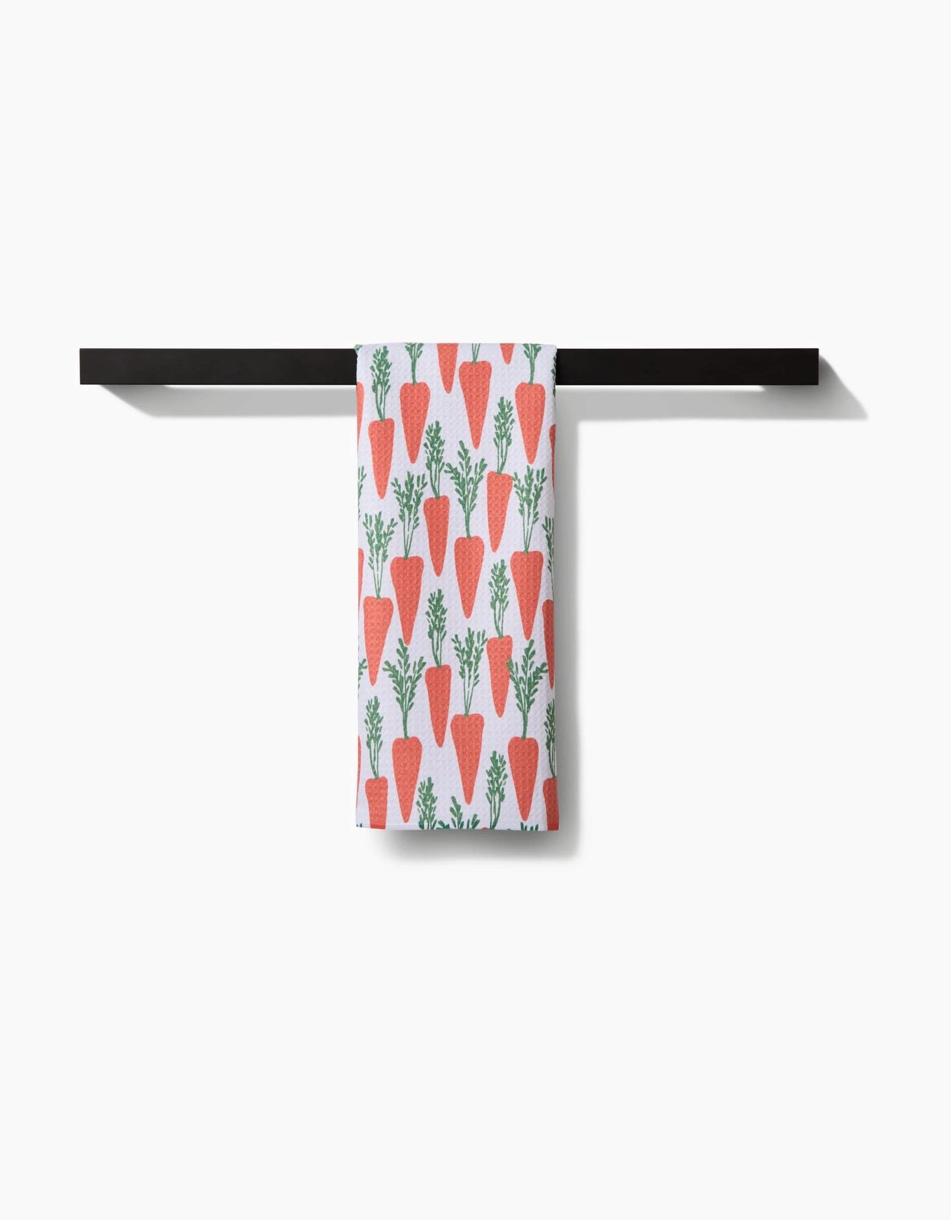 The Spring Carrot Hill tea towel by Hufton Studio, with a red-orange carrot and green top pattern, hangs on a black bar against a white background.