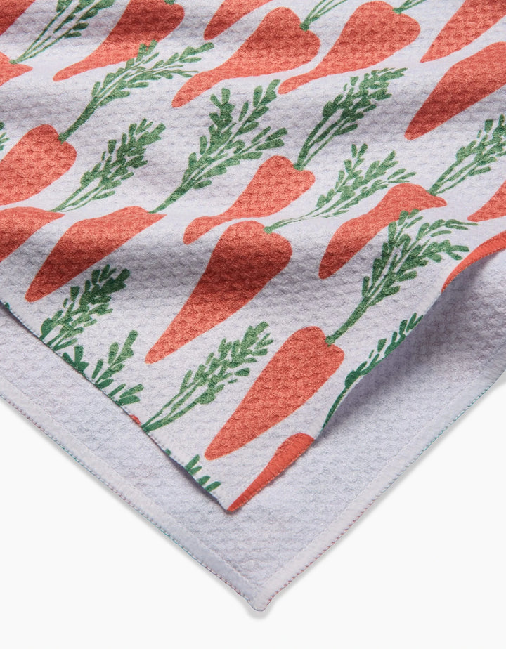 A close-up of the Spring Carrot Hill tea towel by Hufton Studio, showing its white textured fabric with a repeating pattern of orange carrots and green leaves, folded to reveal both the vibrant front and plain underside.