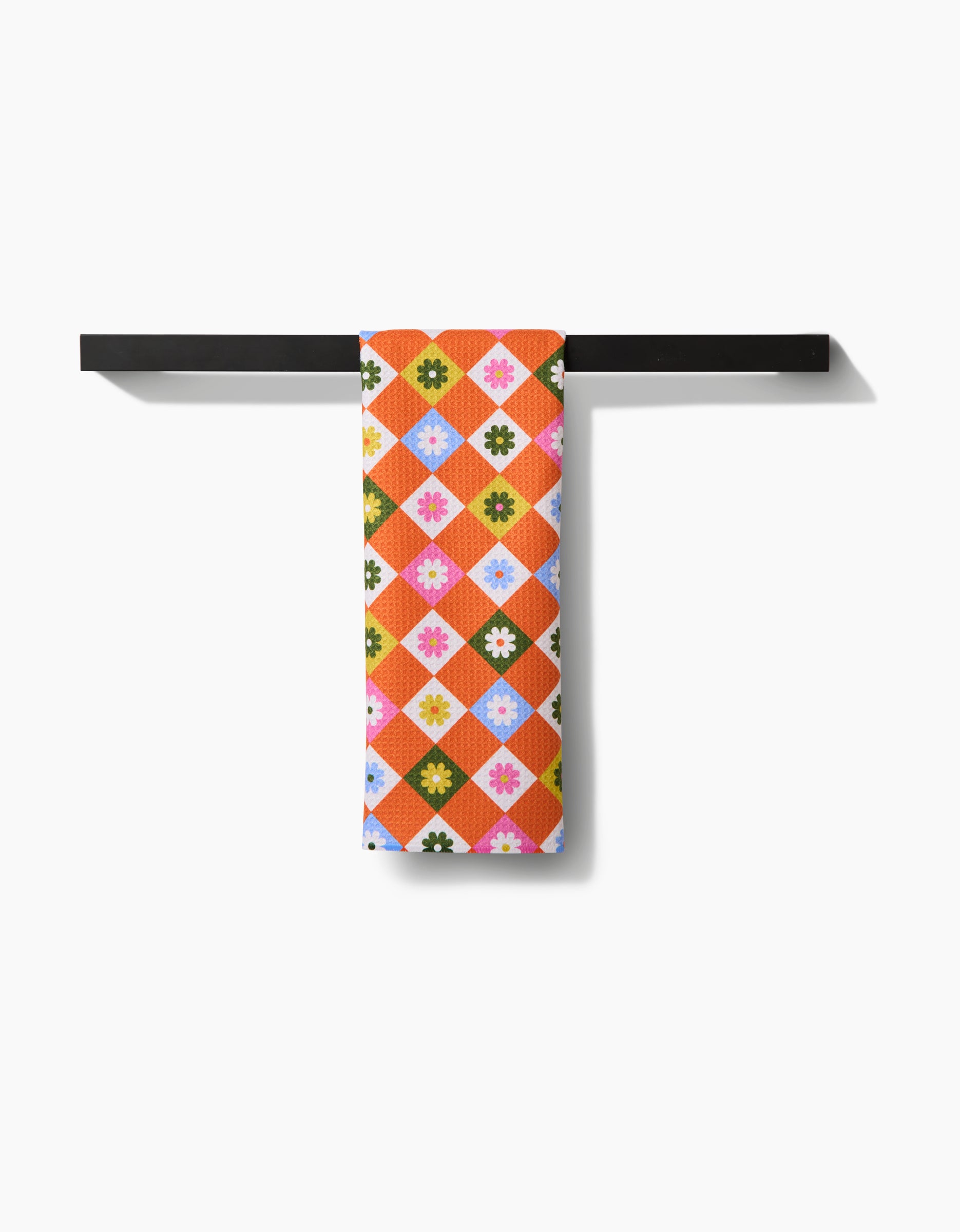 A vibrant Megan Roy Spring Check tea towel with diamond and flower patterns in orange, pink, blue, yellow, and green hangs neatly on a simple black horizontal bar against a white background.