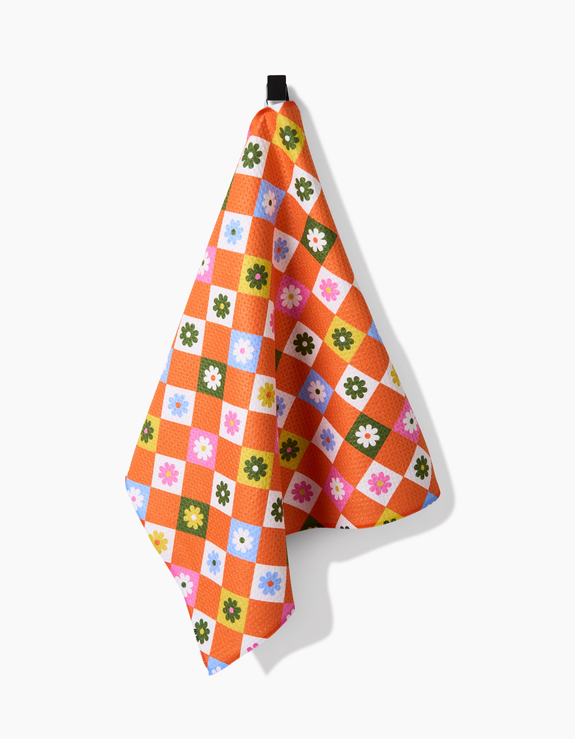 The Megan Roy Spring Check tea towel features a colorful checkered pattern with orange, pink, green, blue, and white squares, each adorned with a small flower. Made from Geoweave fabric for extra absorbency and shown hanging on a hook.