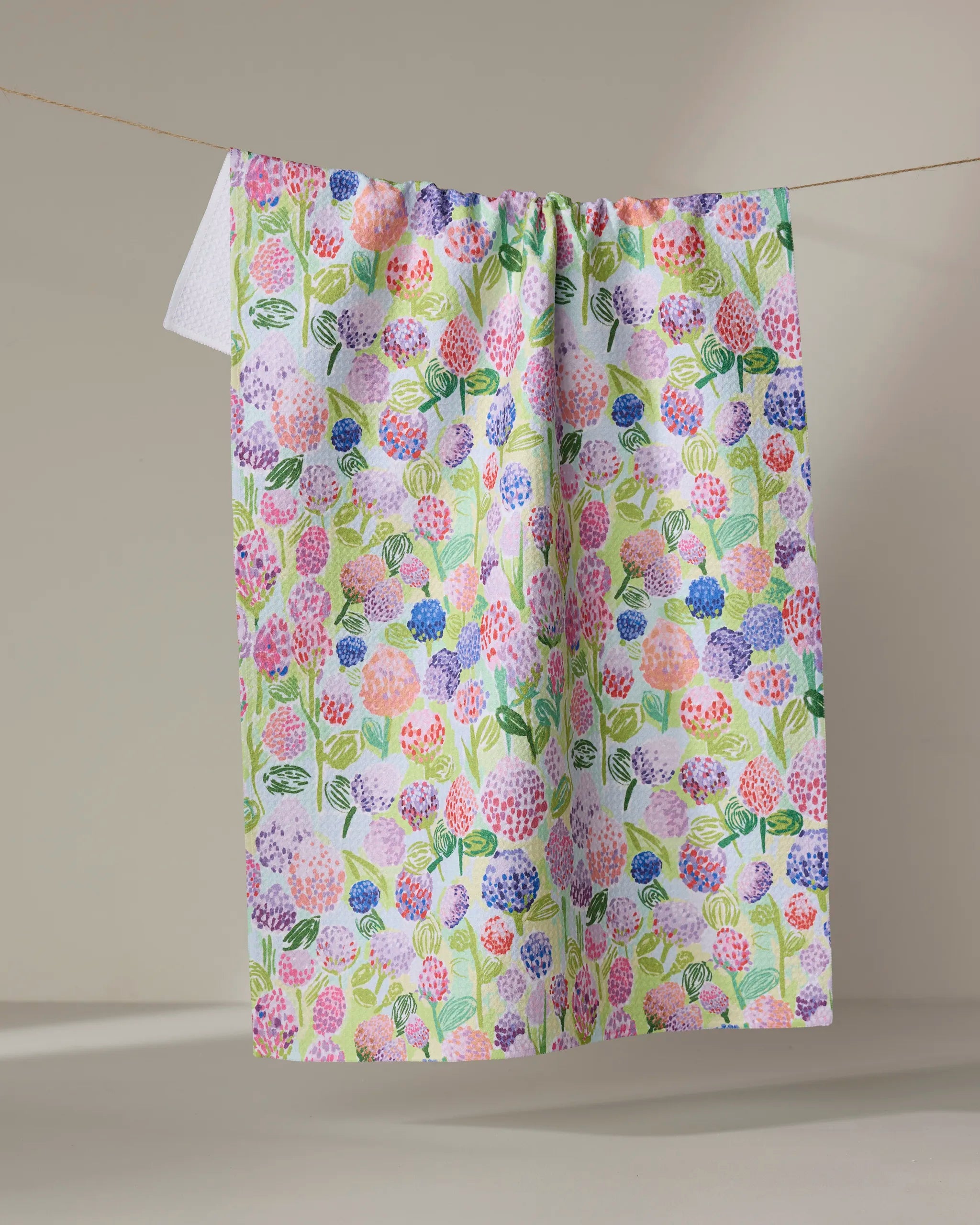 The Spring Clovers kitchen towel by Elisabeth Wing, made from recycled materials, displays pastel flowers and green leaves and hangs on a clothesline against a neutral background.