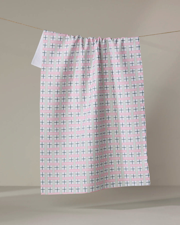 The Spring Cottage Plaid tea towel by Hufton Studio, featuring intersecting pink, blue, and green lines in a grid pattern, hangs on a clothesline against a neutral background.