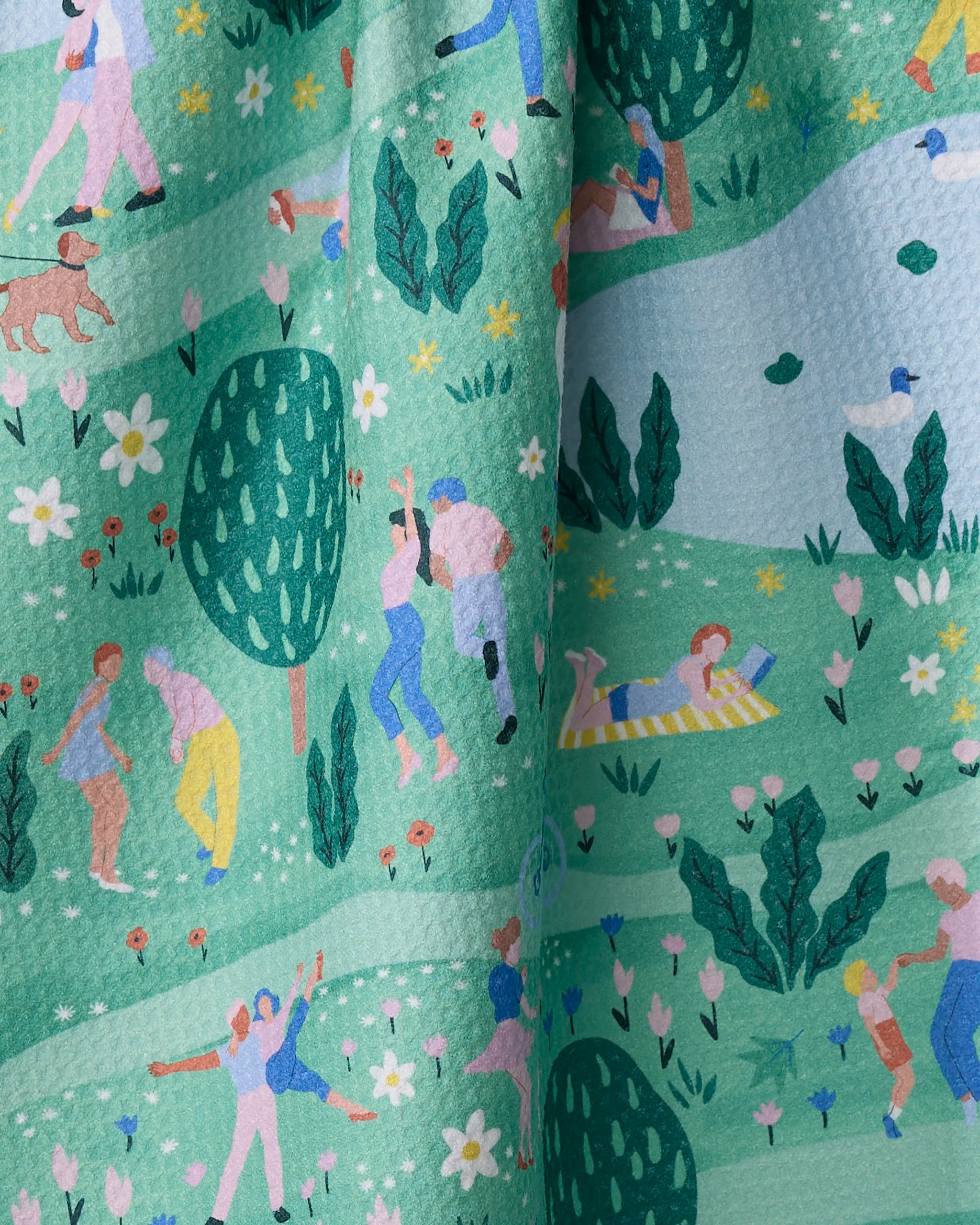The "Spring Festival" quick-drying towel by Ceyda Alasar features illustrations of people celebrating in a vibrant park scene with trees, flowers, ducks, and a dog on a green and blue background.