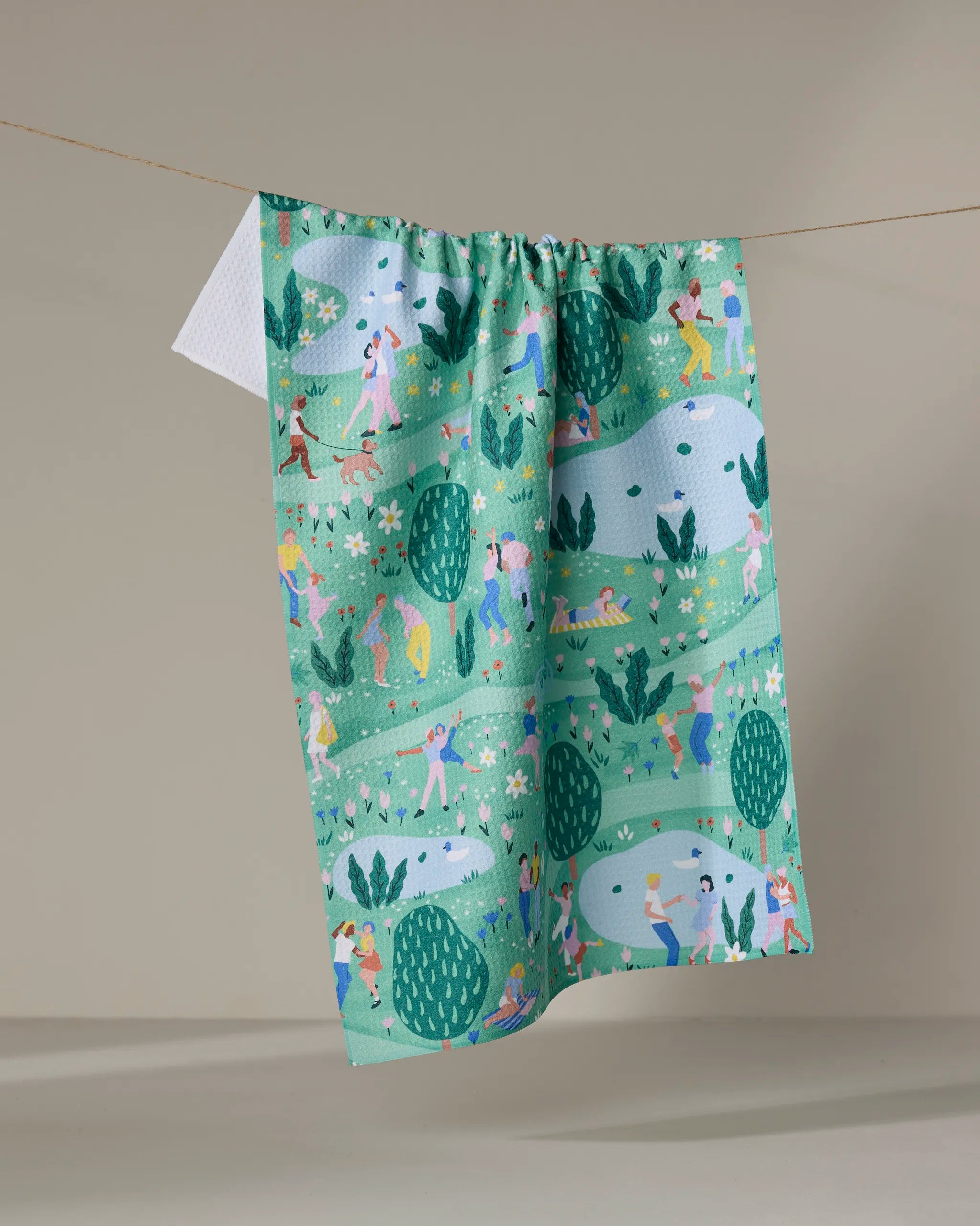 The Spring Festival kitchen towel by Ceyda Alasar features illustrated people walking dogs, biking, jogging, and playing in a park with trees and ponds, all printed on recycled fabric and hung on a string.