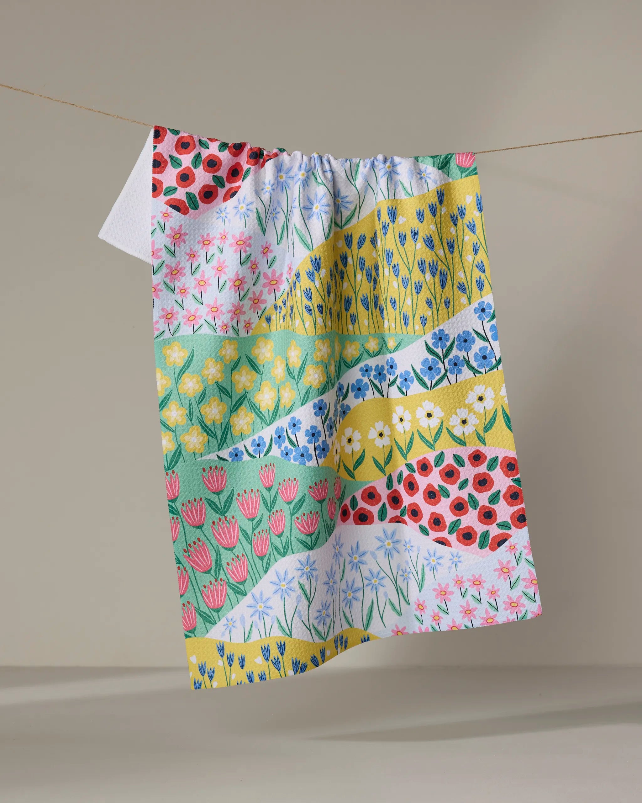 The "Spring Fields" recycled kitchen towel by Ceyda Alasar features vibrant floral patchwork in pink, yellow, green, and blue, hanging from a clothesline against a simple backdrop.